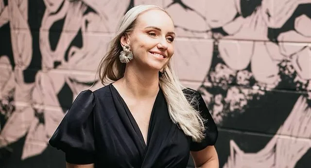 A smiling woman with long blonde hair wearing a black dress and large statement earrings, standing in front of a pink and black abstract mural.