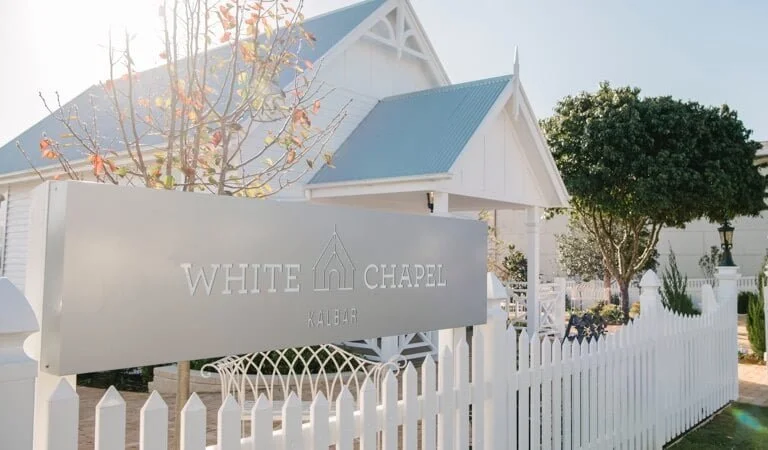 The image shows a white chapel with a blue roof, surrounded by a white picket fence, and a sign that reads "White Chapel Kalbar." There are trees and a garden area around the chapel.