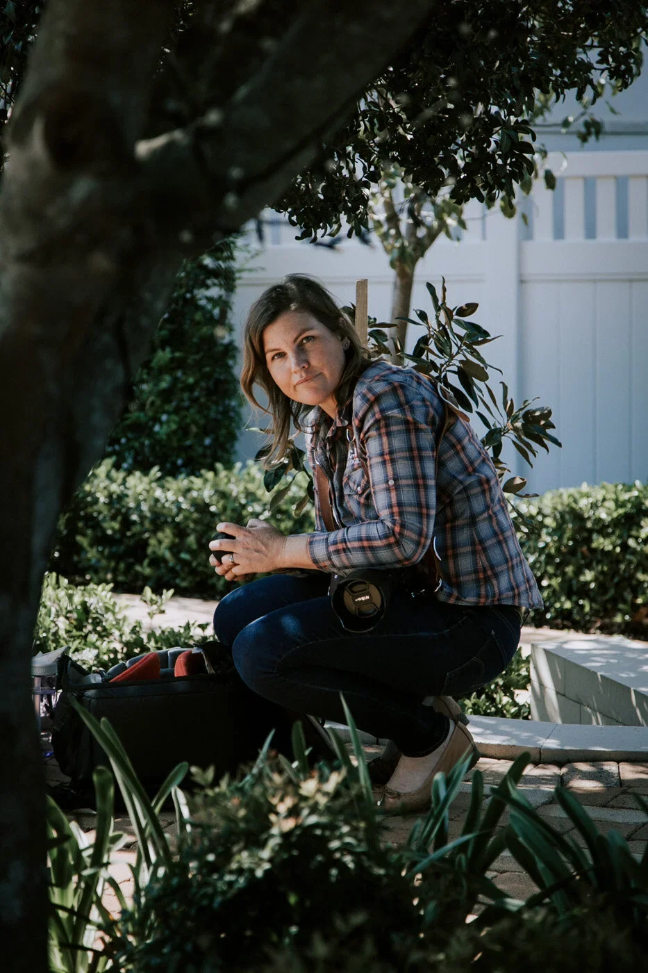 A woman crouches outdoors behind a tree and plants, holding her hands together with a camera hanging from her neck.