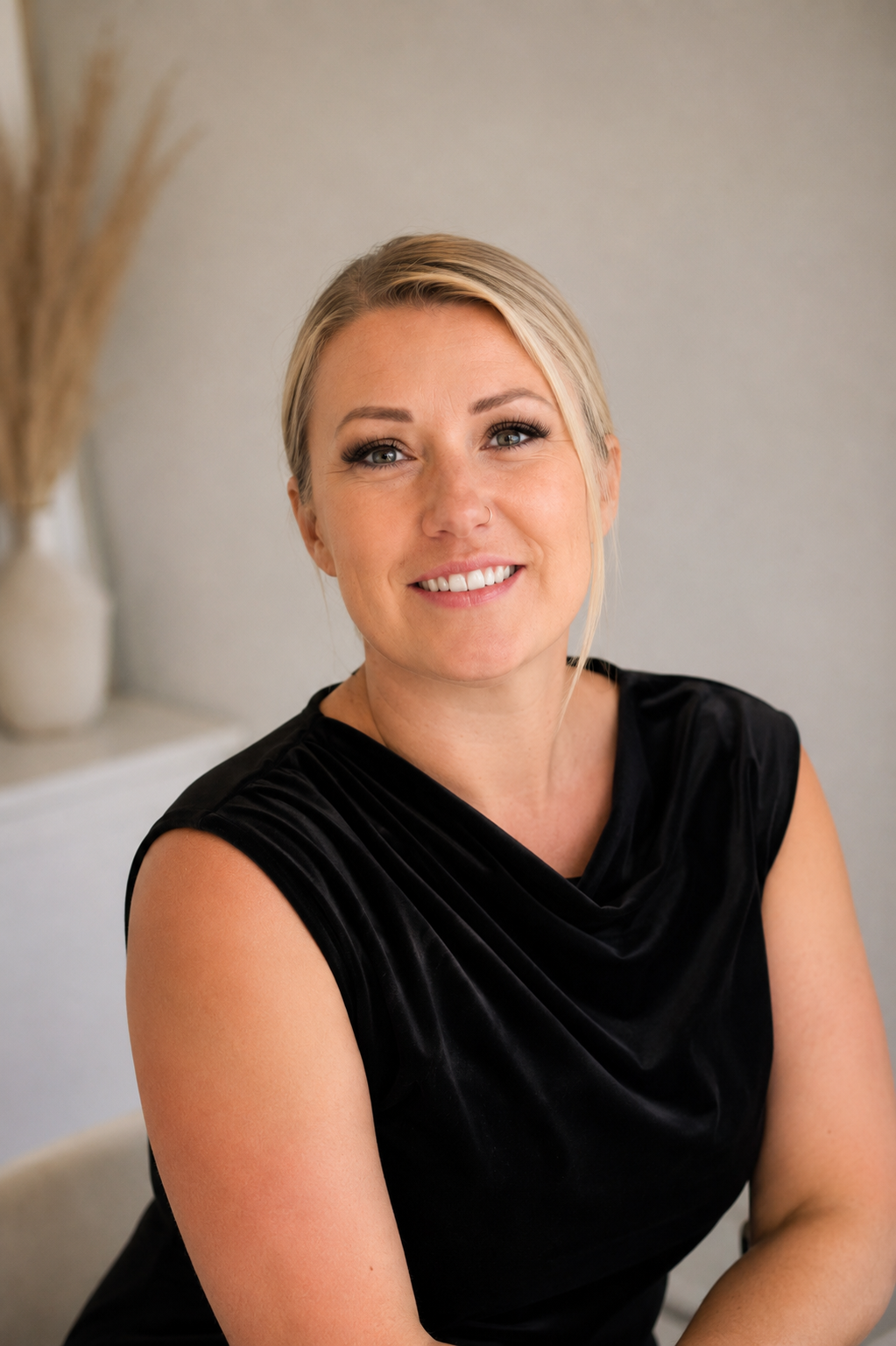 Smiling blonde woman in black sleeveless dress sitting indoors.