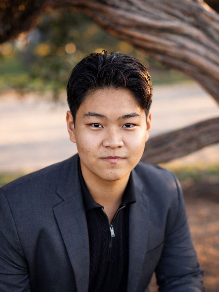 A young man in a dark suit jacket and zip-up sweater sitting outdoors near a tree, with blurred trees and landscape in the background.