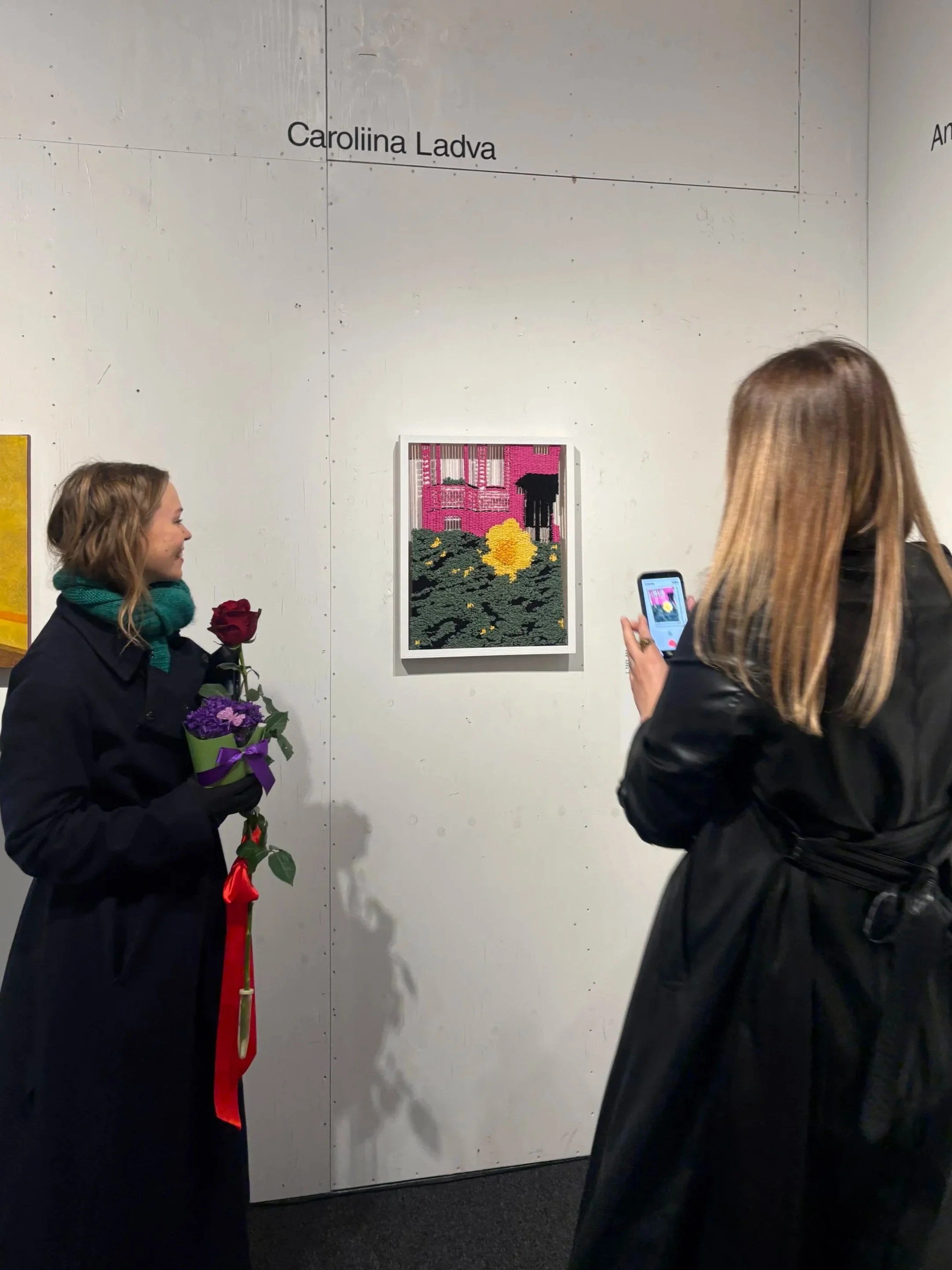Two women are standing in front of a wall with names of artists, looking at a piece of artwork. One woman, holding a rose and a small bouquet, is smiling. The other woman, taking a photo with her phone, is facing the artwork. The artwork is art piece of a pink building with greenery and a yellow sun.