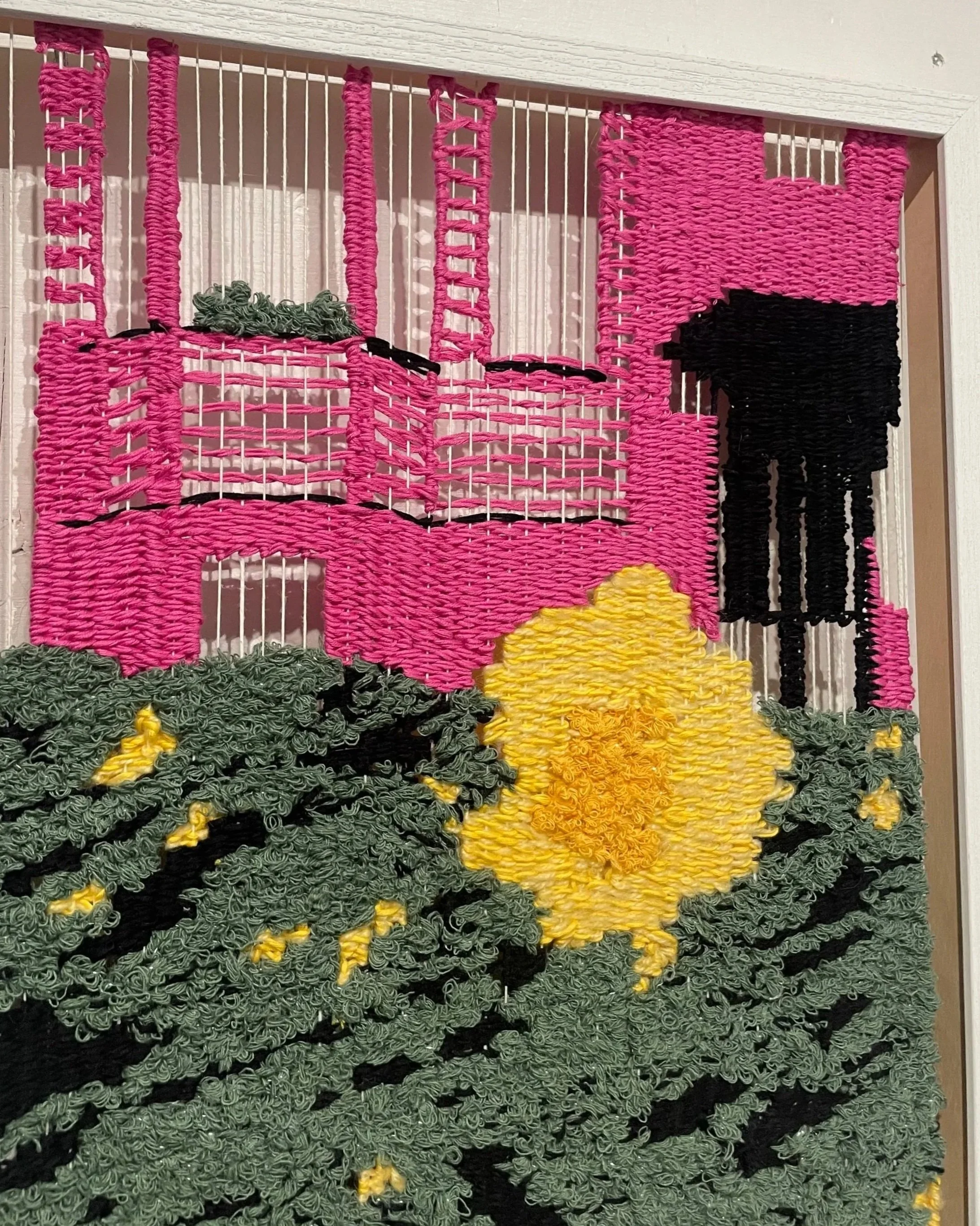 A colorful woven wall hanging depicting  a pink house with a black door, surrounded by green grass and a yellow flower, made from yarn and string.