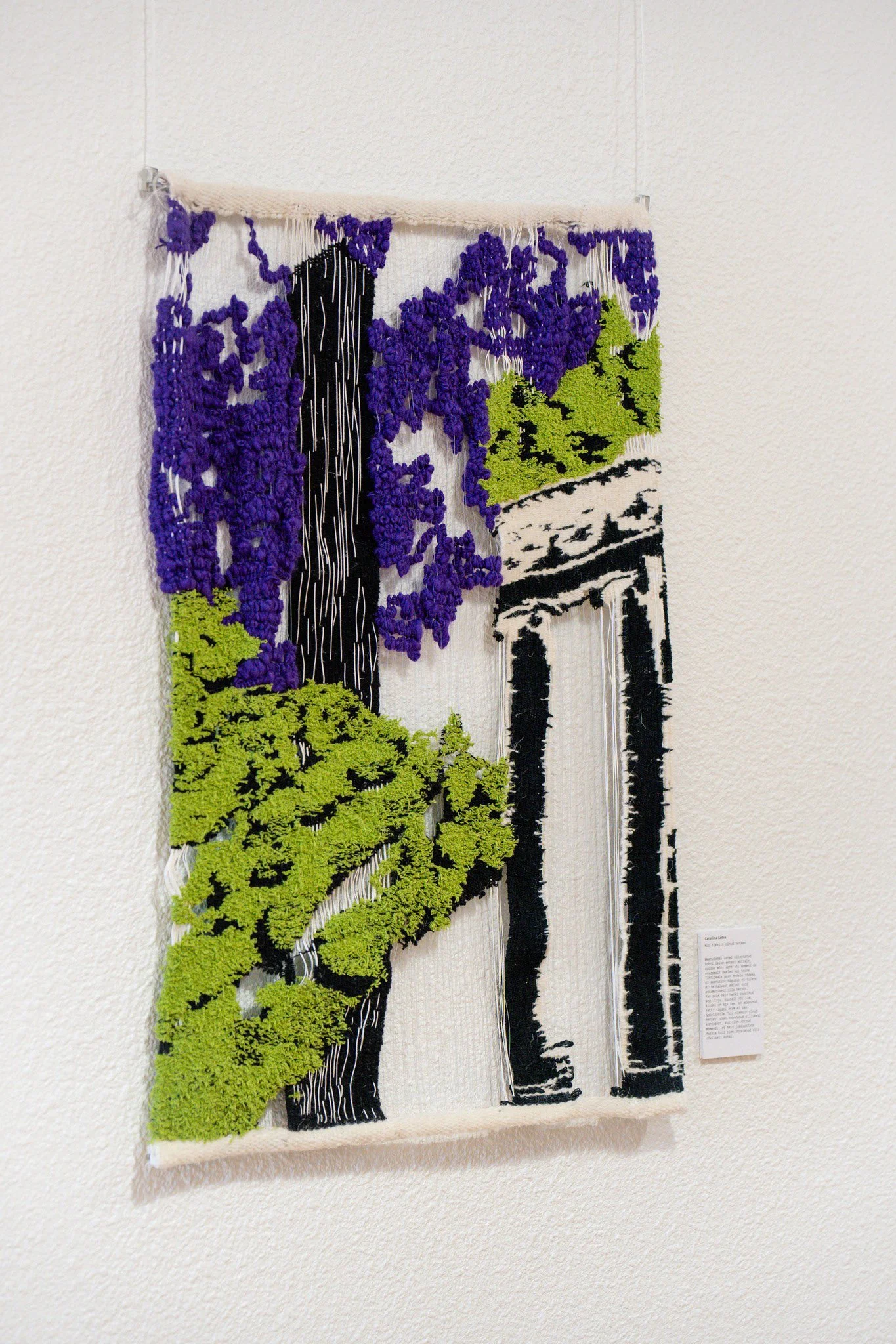 Textile art piece depicting a landscape with purple and green elements, and a black and white building in the background, hung on a white wall.