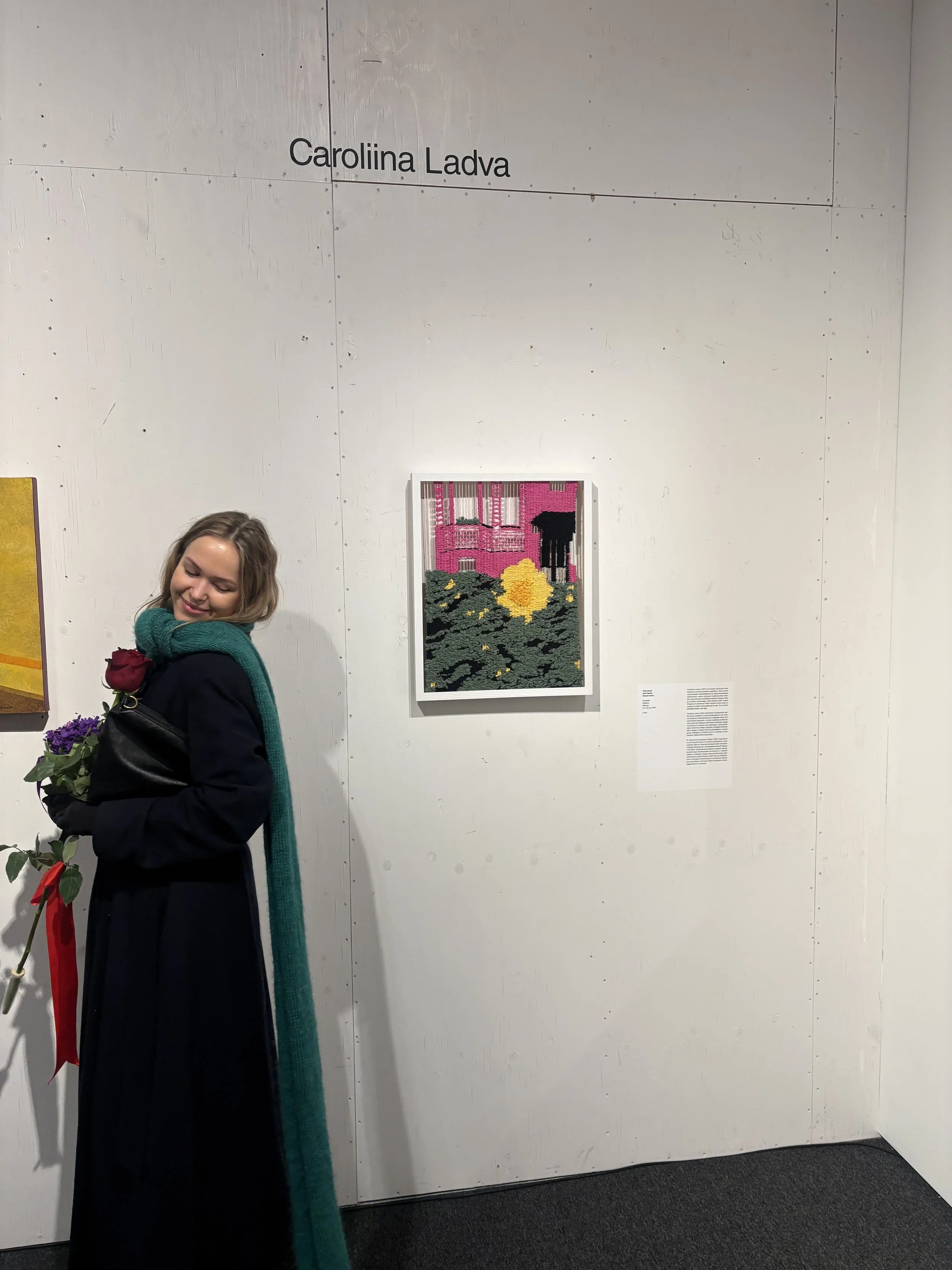 A woman with shoulder-length hair smiling, holding a bouquet of purple flowers and a single red rose, standing in front of a white wall at an art exhibit. The wall features a framed artwork of a pink building with a  yellow flower in the foreground. The name 'Carolina Ladva' is displayed above the artwork.