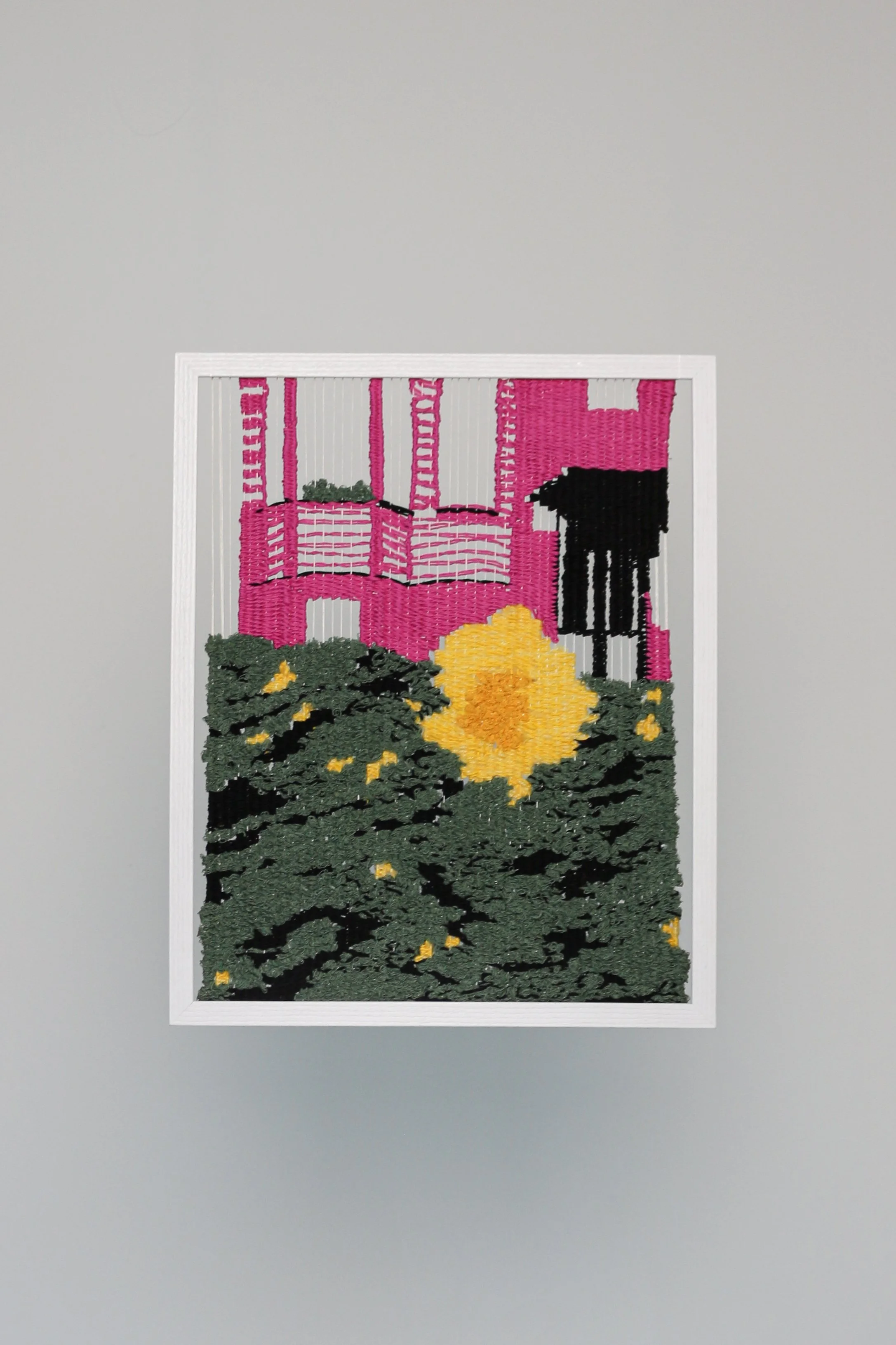 Woven textile artwork displaying a pink house like shape, green abstract landscape and a yellow flower in the foreground, mounted on a white wall.