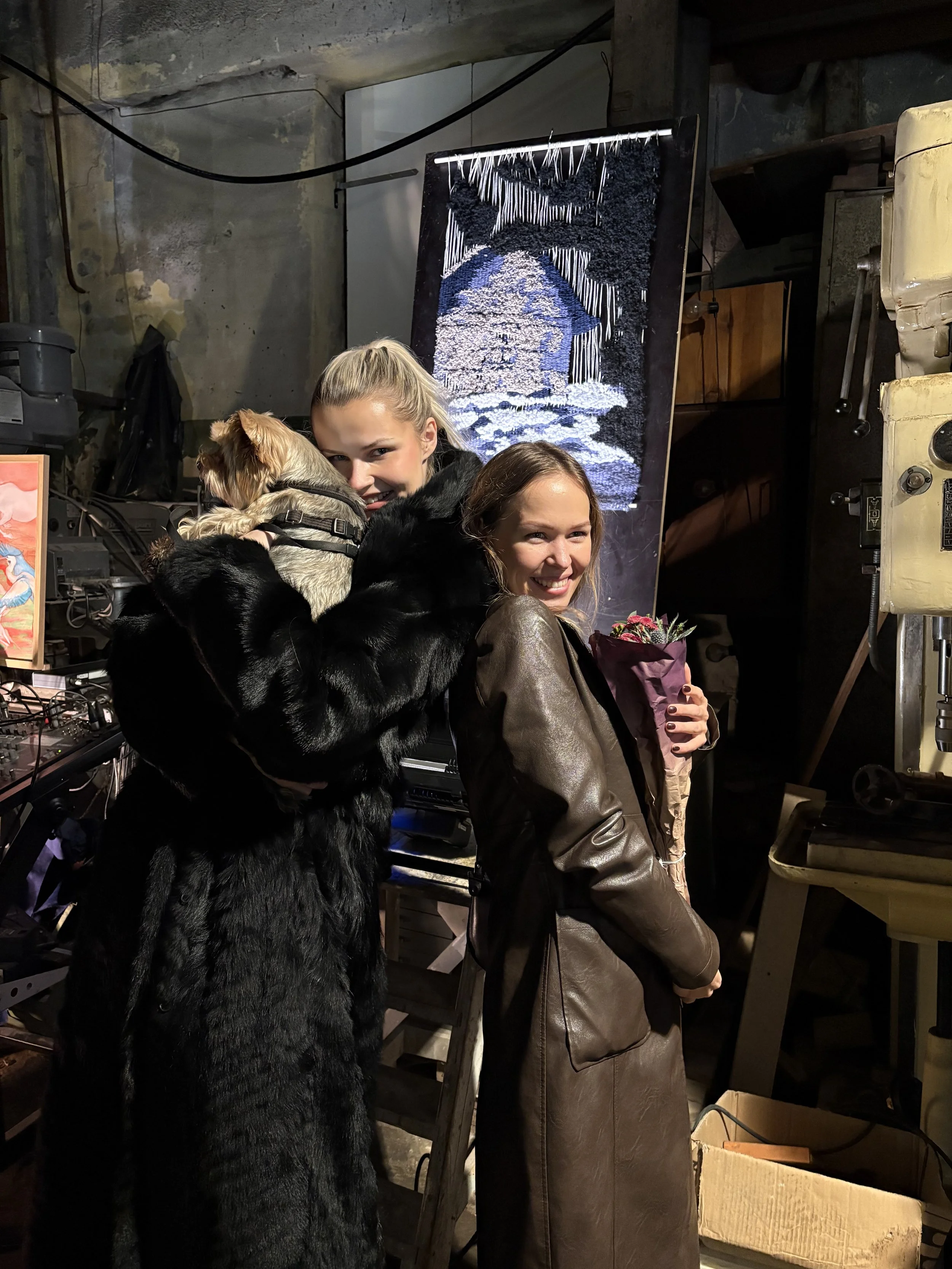 Two women are smiling, one holding a bouquet of flowers, while a dog is being hugged by the woman in the background in an industrial setting with a textile art piece in the background.
