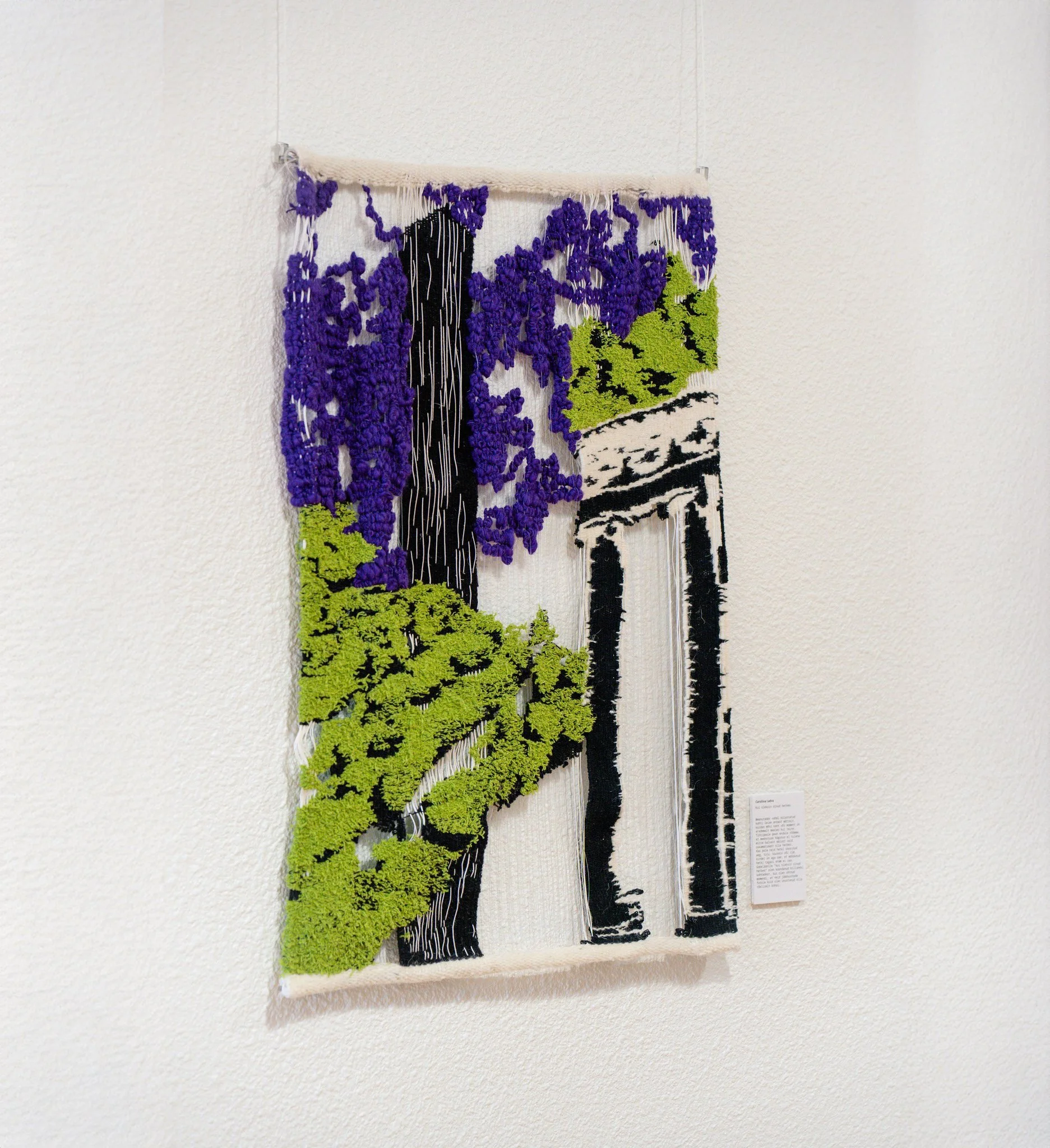 Textile wall hanging art featuring a landscape, skyscraper, purple sky, green foliage, and black and white design elements, mounted on a white wall.