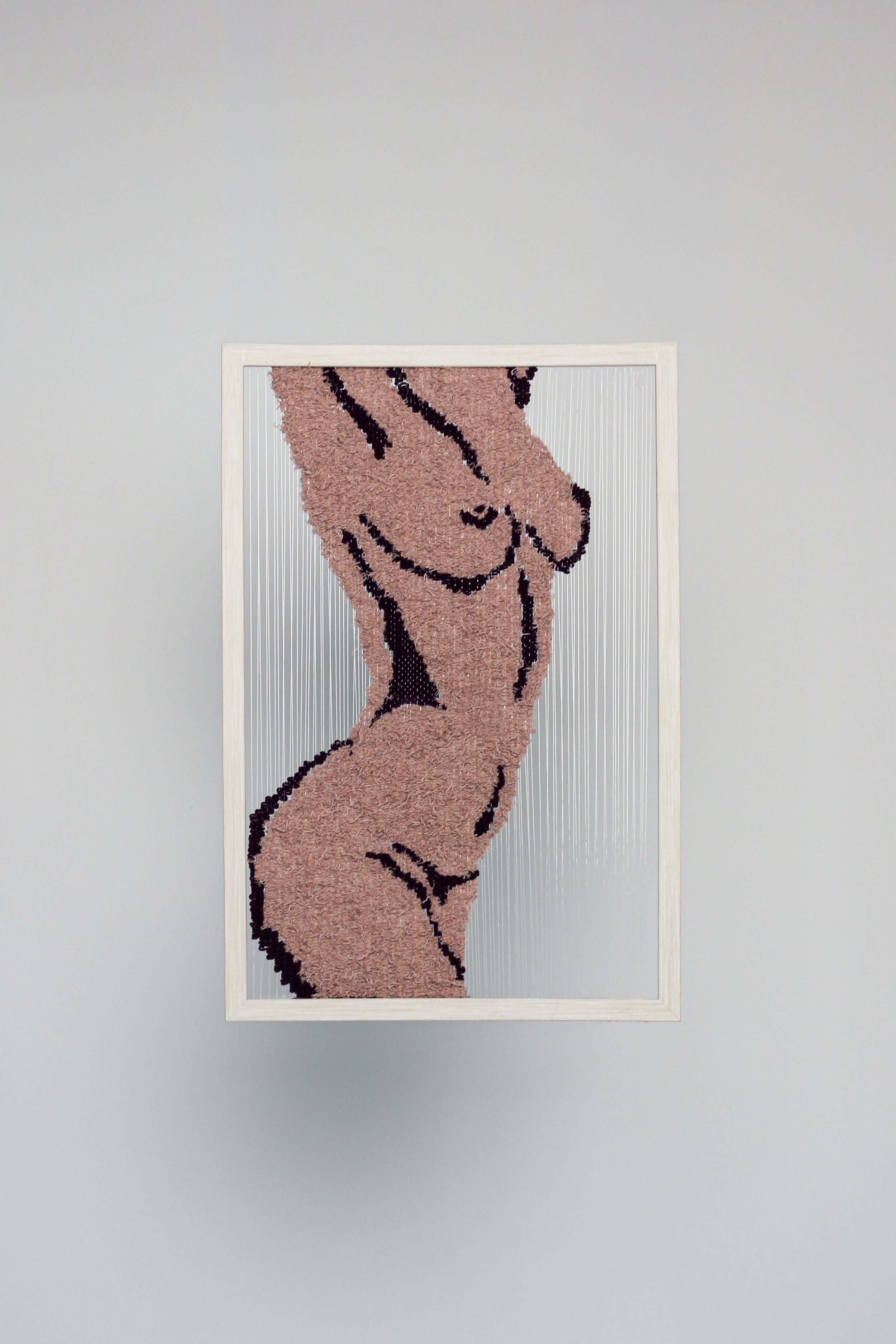 Framed textile art of a stylized nude female figure, displayed on a light gray wall.
