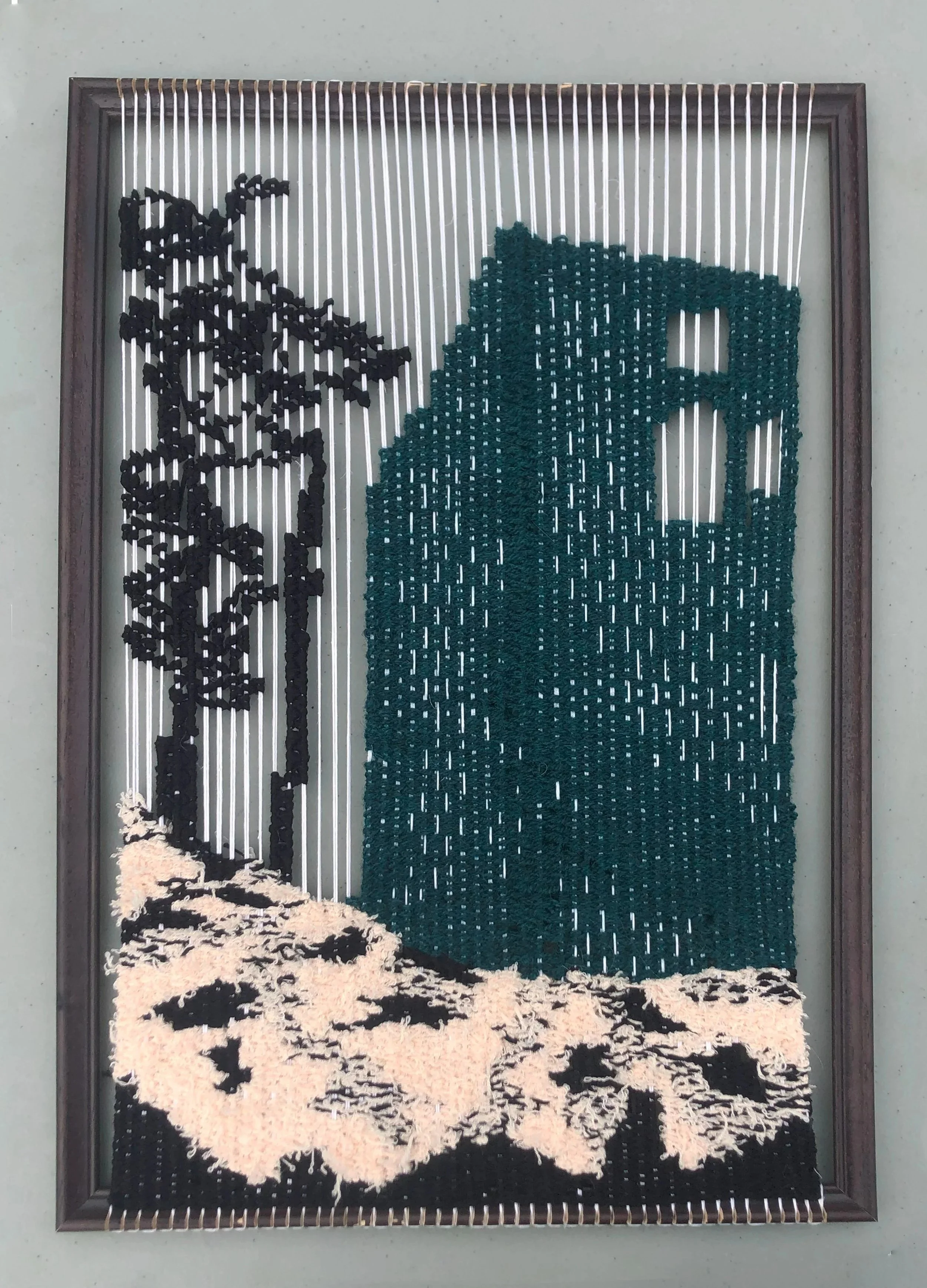 Textile art piece with black, teal, and beige threads on a frame, depicting a landscape, trees and a large building.