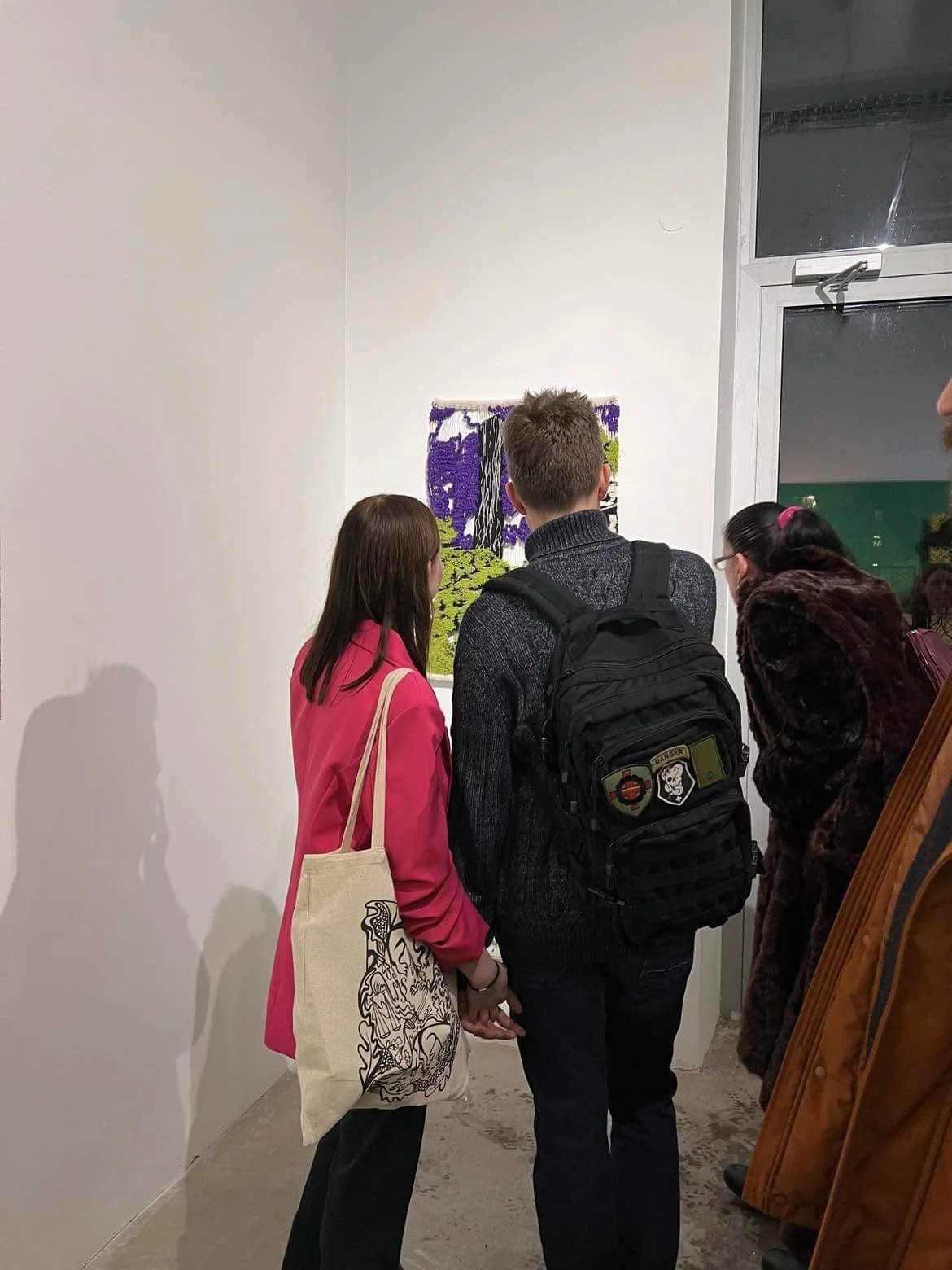 People standing in a gallery observing a piece of artwork on a white wall. The artwork appears to be a fabric or textile piece with a landscape scene, showing trees and grass in bright colors.
