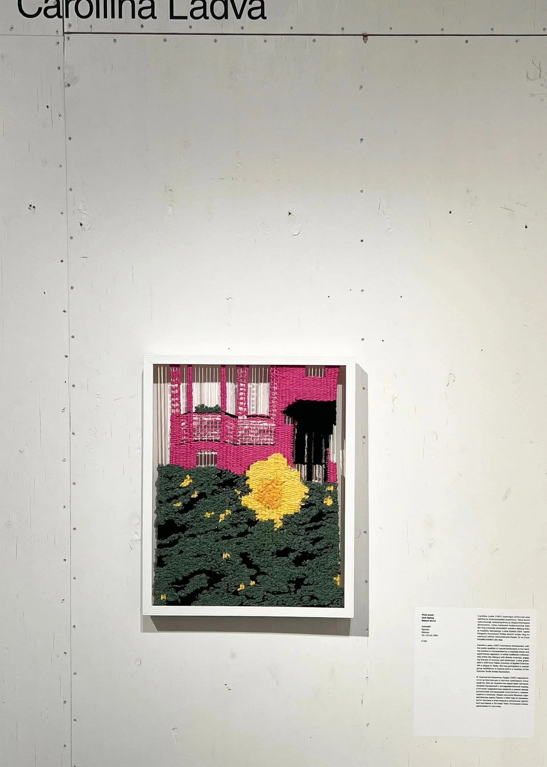 A framed textile art piece depicting a vibrant landscape scene with pink house, lush green landscape and a large yellow flower displayed on a white wall.