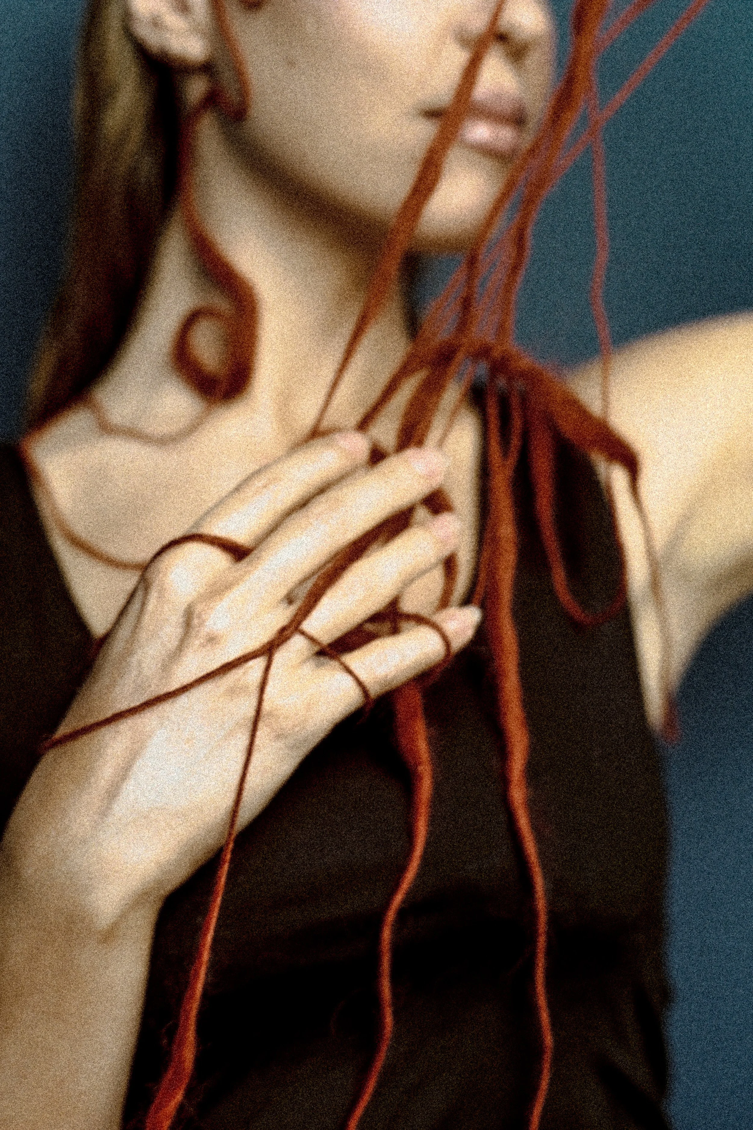 A close-up of a woman with reddish-brown decorative yarns over her, touching her face and shoulder, with a blurred dark background.