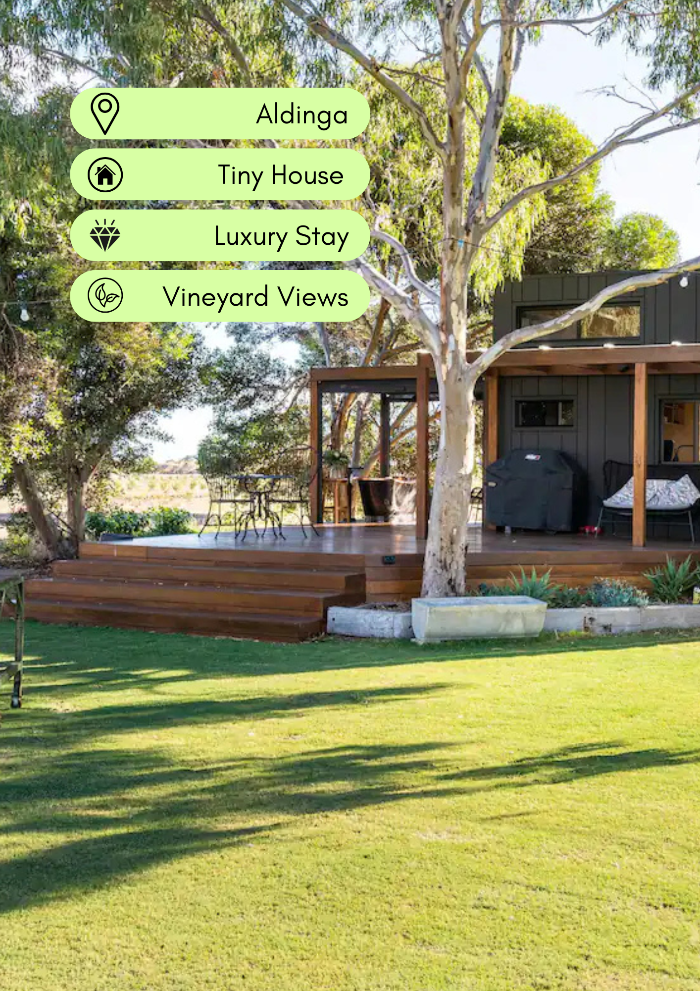 A backyard scene with a raised wooden deck, patio furniture, a barbecue grill, and a modern house with large window and a wooden frame, surrounded by green grass and trees, during daylight.