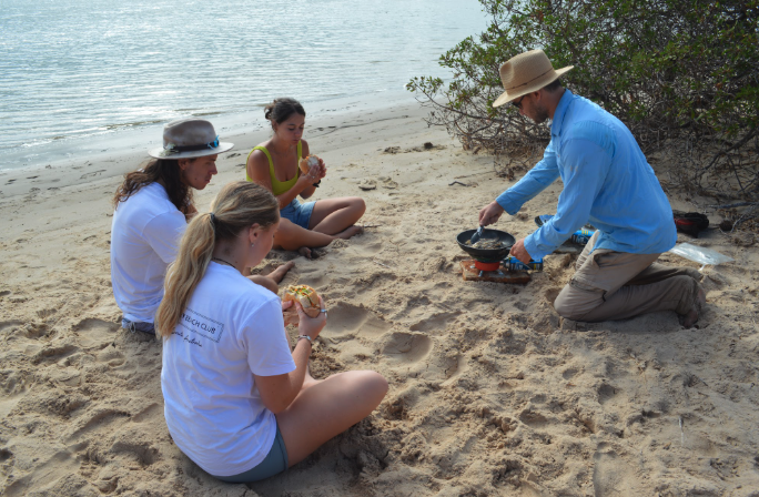 Four young women with long hair sitting on sandy beach, two holding sandwiches, watching man cooking food in frying pan over camp stove near shrubs.