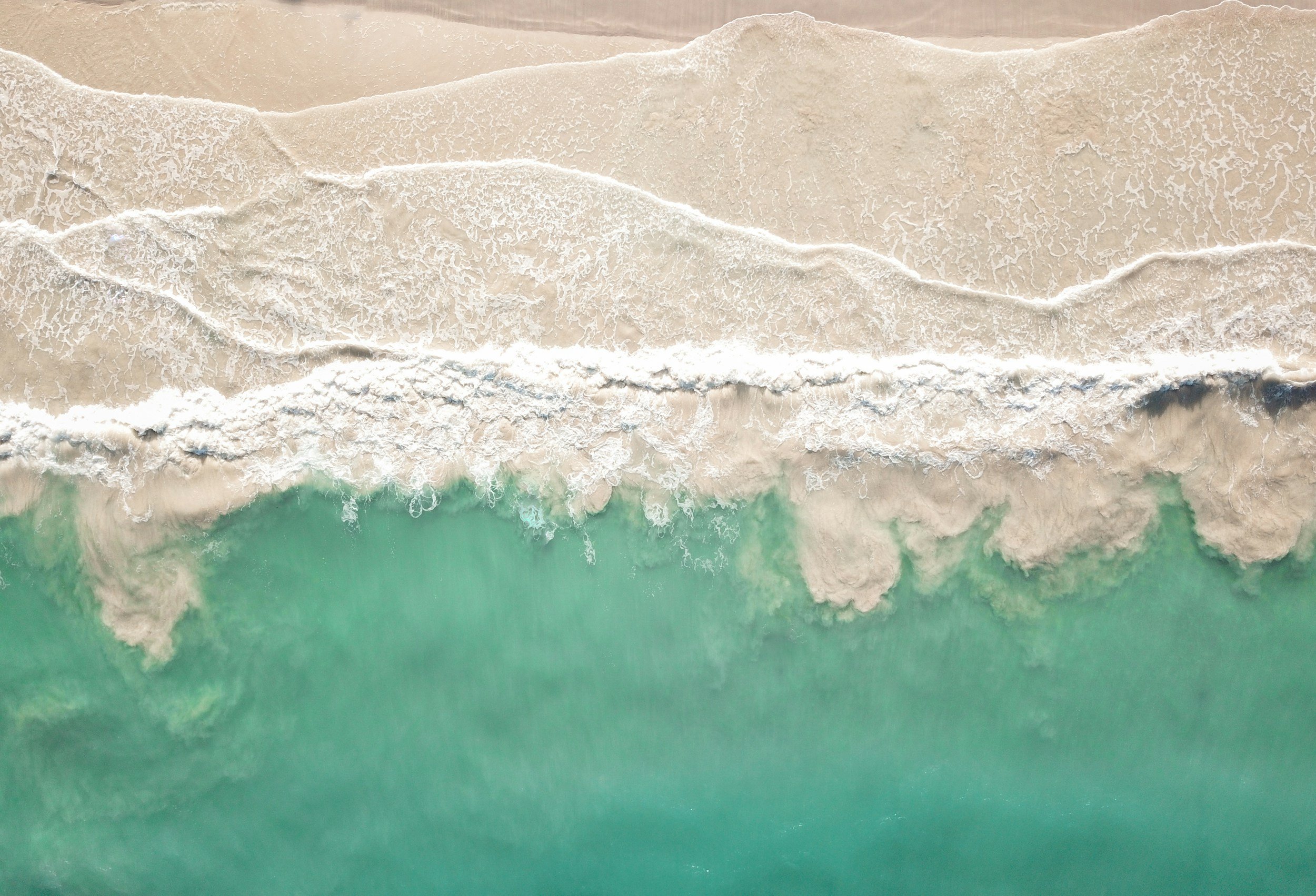 Aerial view of shoreline with waves crashing onto sandy beach with clear turquoise water.