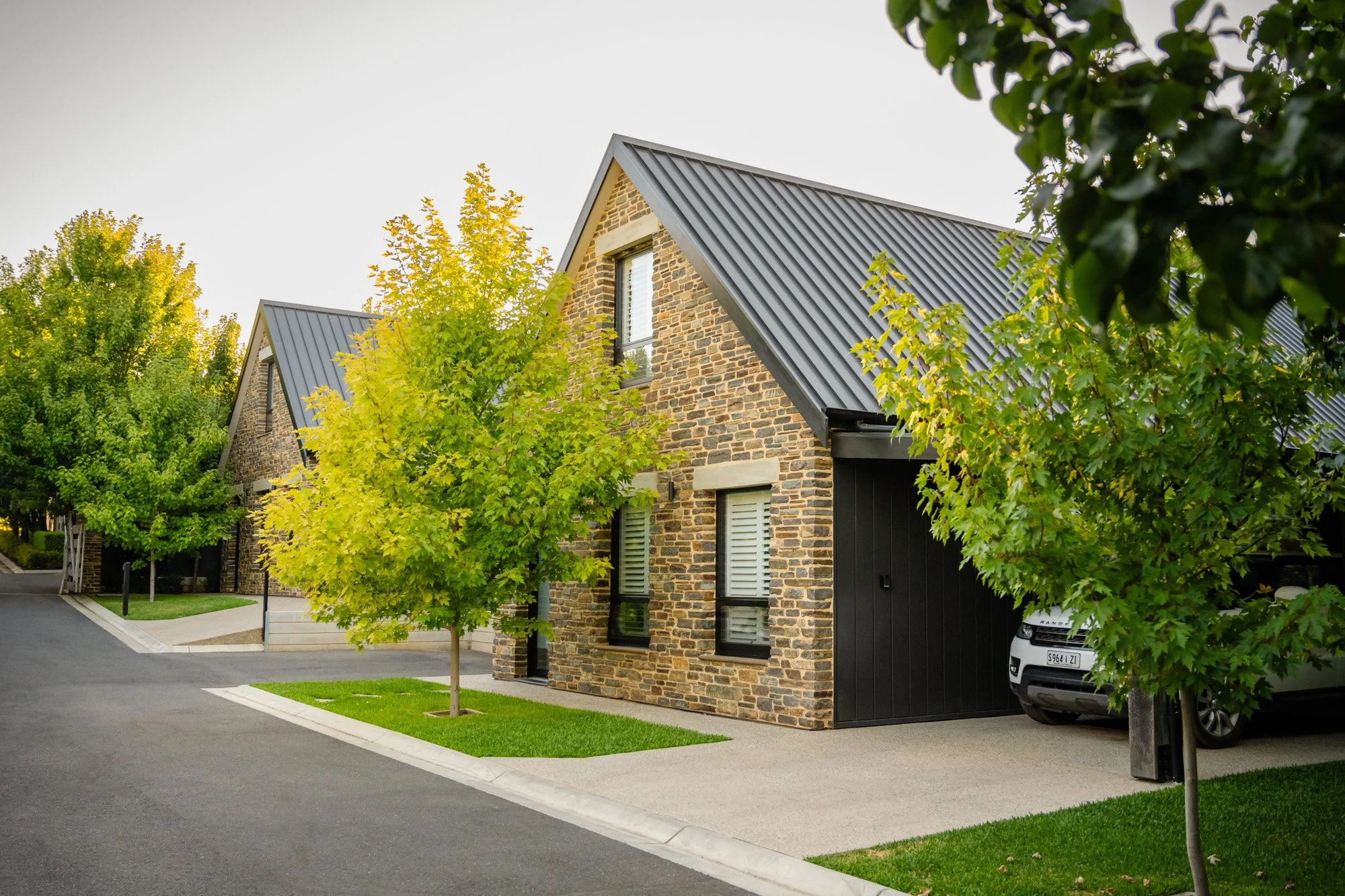 A modern house with brick exterior and a dark metal roof, surrounded by green trees and a well-maintained lawn in a suburban neighborhood.
