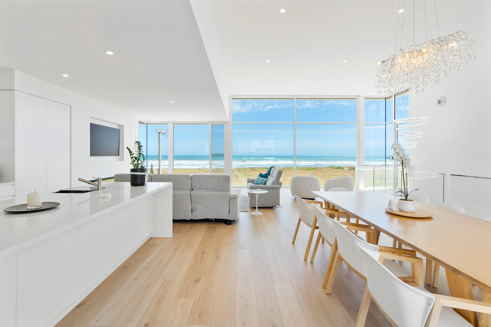 Modern living room with large windows overlooking the ocean, white walls, wooden floors, a white kitchen counter, a gray sofa, white dining chairs, and decorative lighting fixtures.