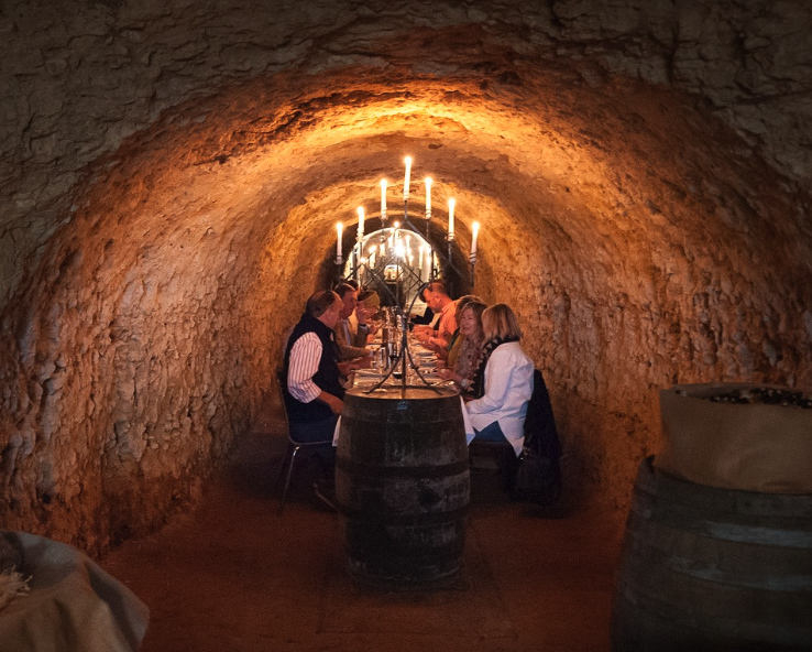 People dining at a long table inside a cave with lit candles and wine barrels.