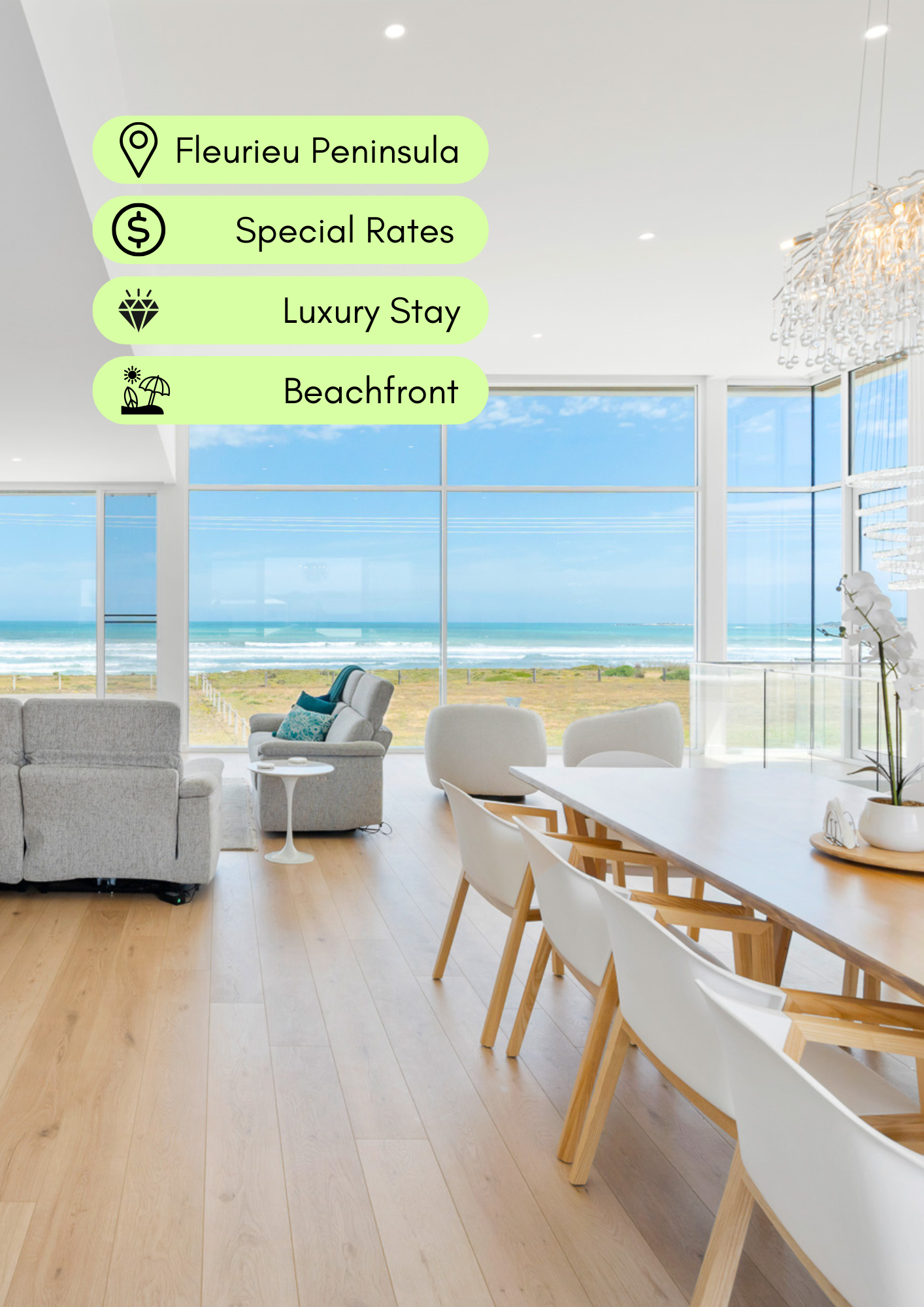 Bright, modern beachfront interior with large windows showing ocean view, white and light wood furniture, and a living room with sofas, along with a dining area with white and wooden chairs.