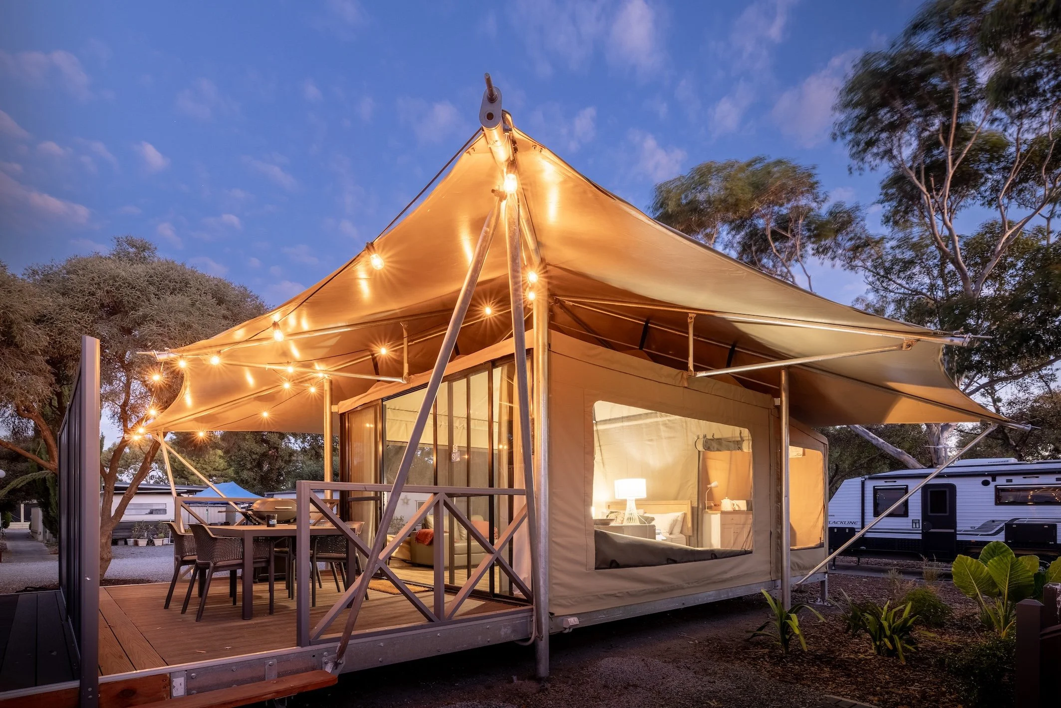 A cozy outdoor living space with a large beige canopy tent decorated with string lights. Inside, there are chairs, a table, and a lamp, creating a warm ambiance. Trees and caravans are visible in the background during twilight.