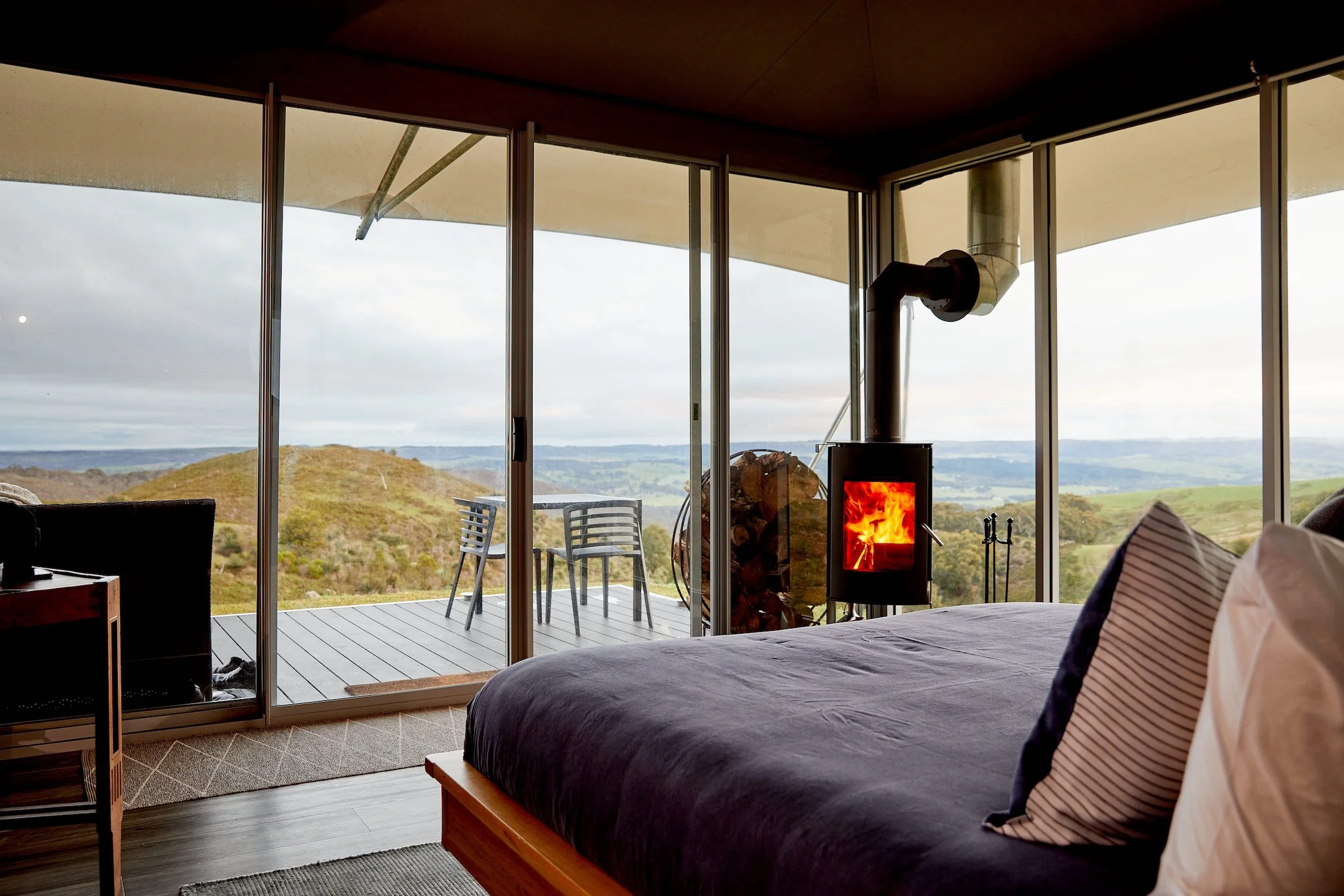 Interior of a modern bedroom with large glass sliding doors opening to a deck with outdoor chairs and a wood stove, overlooking rolling hills and cloudy sky.
