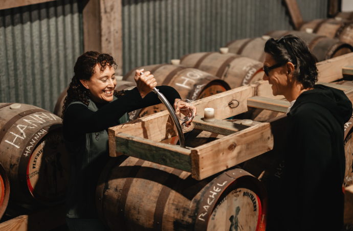 Two women with dark hair, one with glasses, laughing and pouring whiskey into a glass from a spout, in a warehouse with whiskey barrels.
