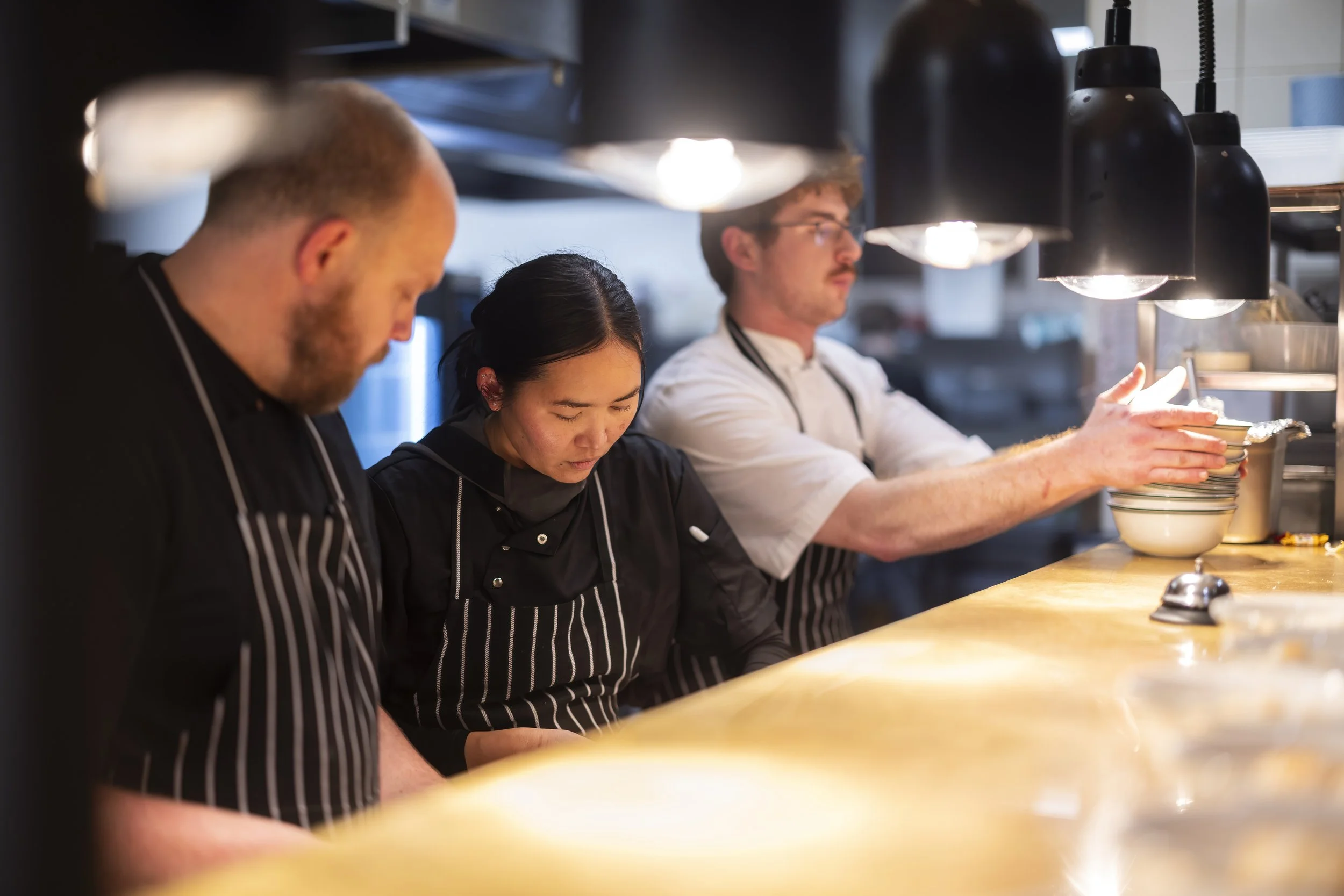 Three chefs working at a kitchen counter, focused on their tasks, with black lamps overhead illuminating their workspace.