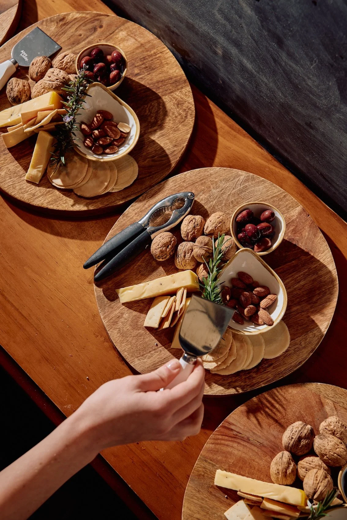 Two wooden platters with assorted cheeses, walnuts, almonds, cherries, and garnished with sprigs of rosemary, served on a wooden table, with a hand holding a cheese slicer.