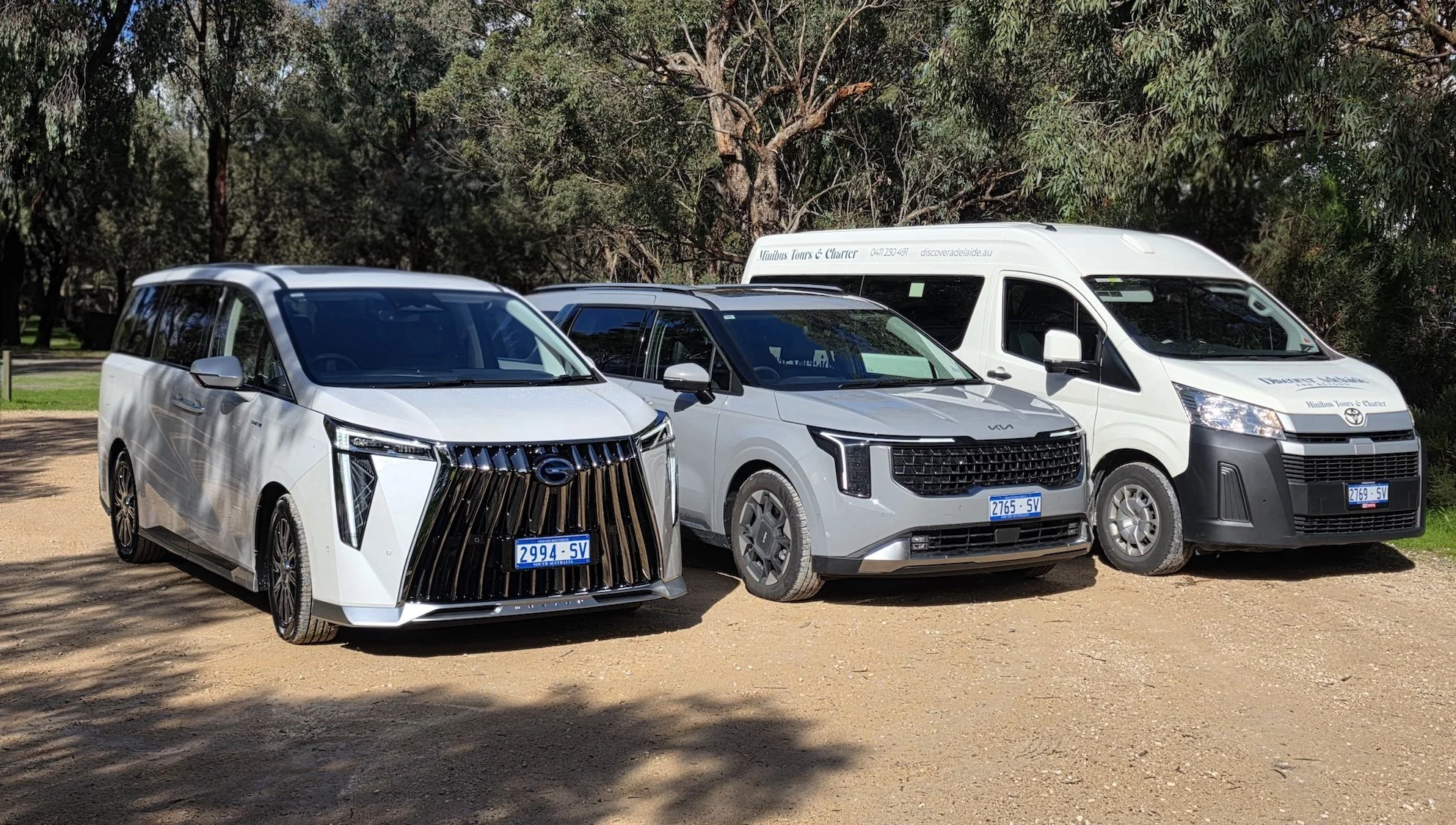 Three vehicles parked on a dirt area with trees in the background. The cars include a white van and a gray SUV, and what appears to be another gray van. The vehicles are parked side by side, facing the same direction.