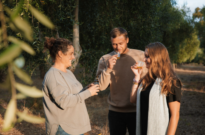 Three friends smiling and clinking glasses of beer outdoors in a wooded area on a sunny day.