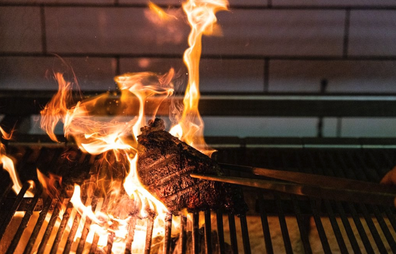 A piece of steak grilling on a barbecue with flames around it.