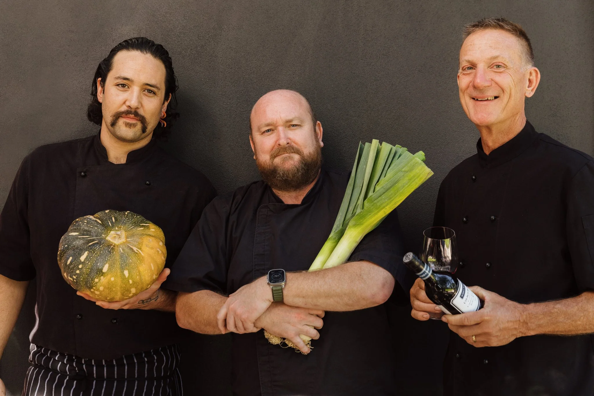 Group of three male chefs standing against a dark wall, holding vegetables and wine, smiling.