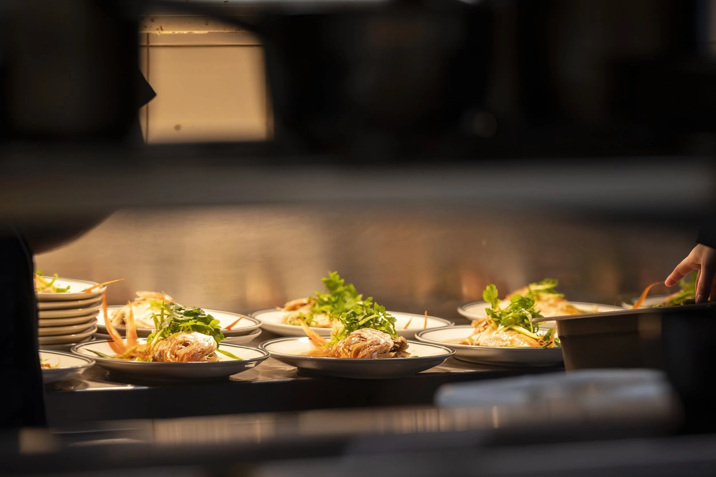 A view through a kitchen serving window showing plates of prepared food, including seafood garnished with greens, on a counter inside a restaurant.