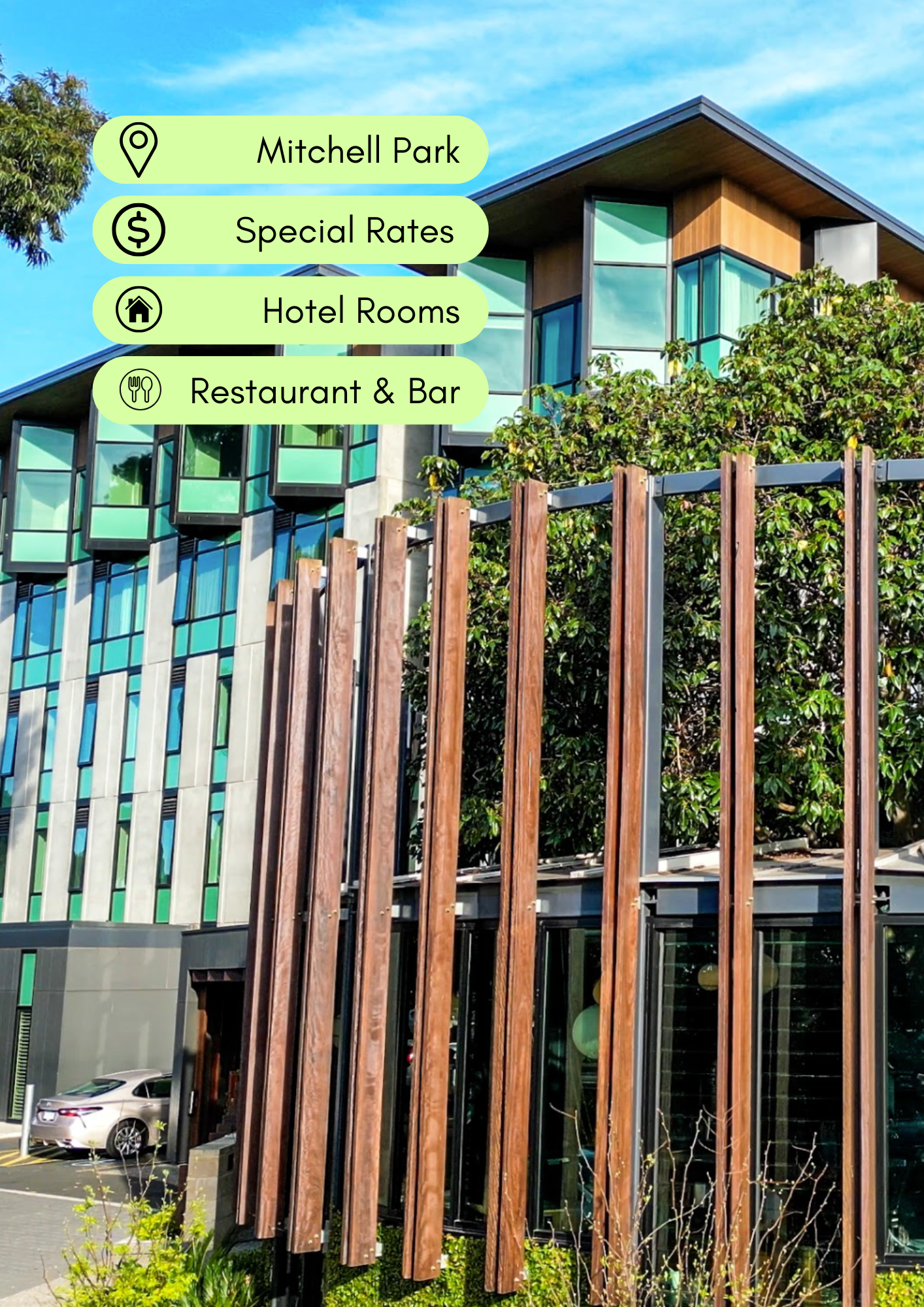 Exterior view of a modern building with green-tinted glass windows, vertical wooden slats on the facade, greenery in the foreground, and a parking lot with a white car. Clear blue sky overhead.
