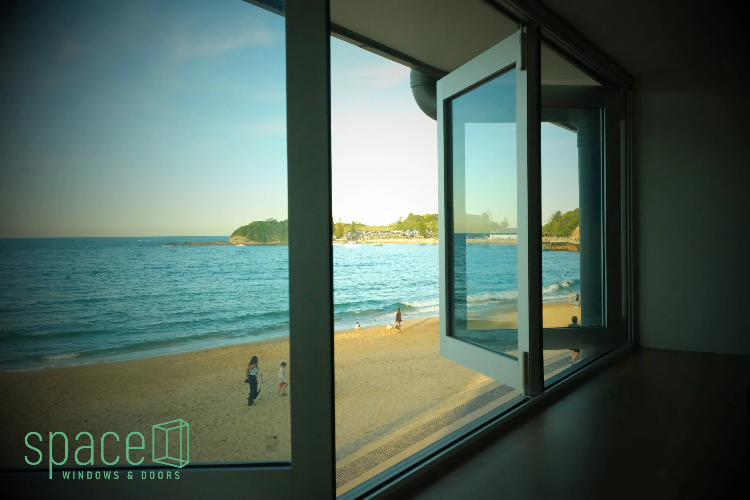 View of a beach through a large open window with a logo of 'spacel WINDOWS & DOORS' in the bottom-left corner.
