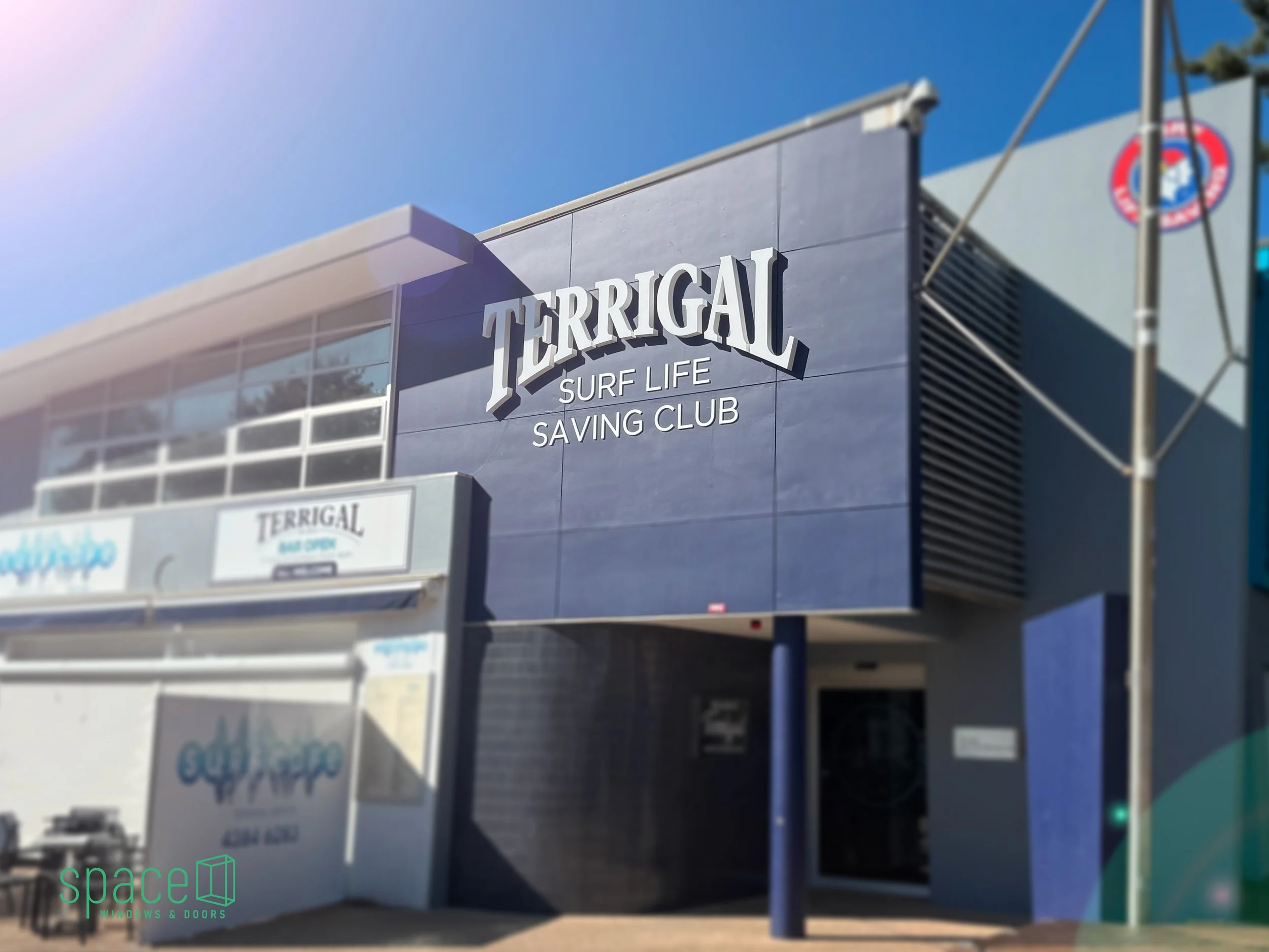 Exterior of a modern building with a sign that reads 'TERRIGAL SURF LIFE SAVING CLUB' under a clear blue sky.