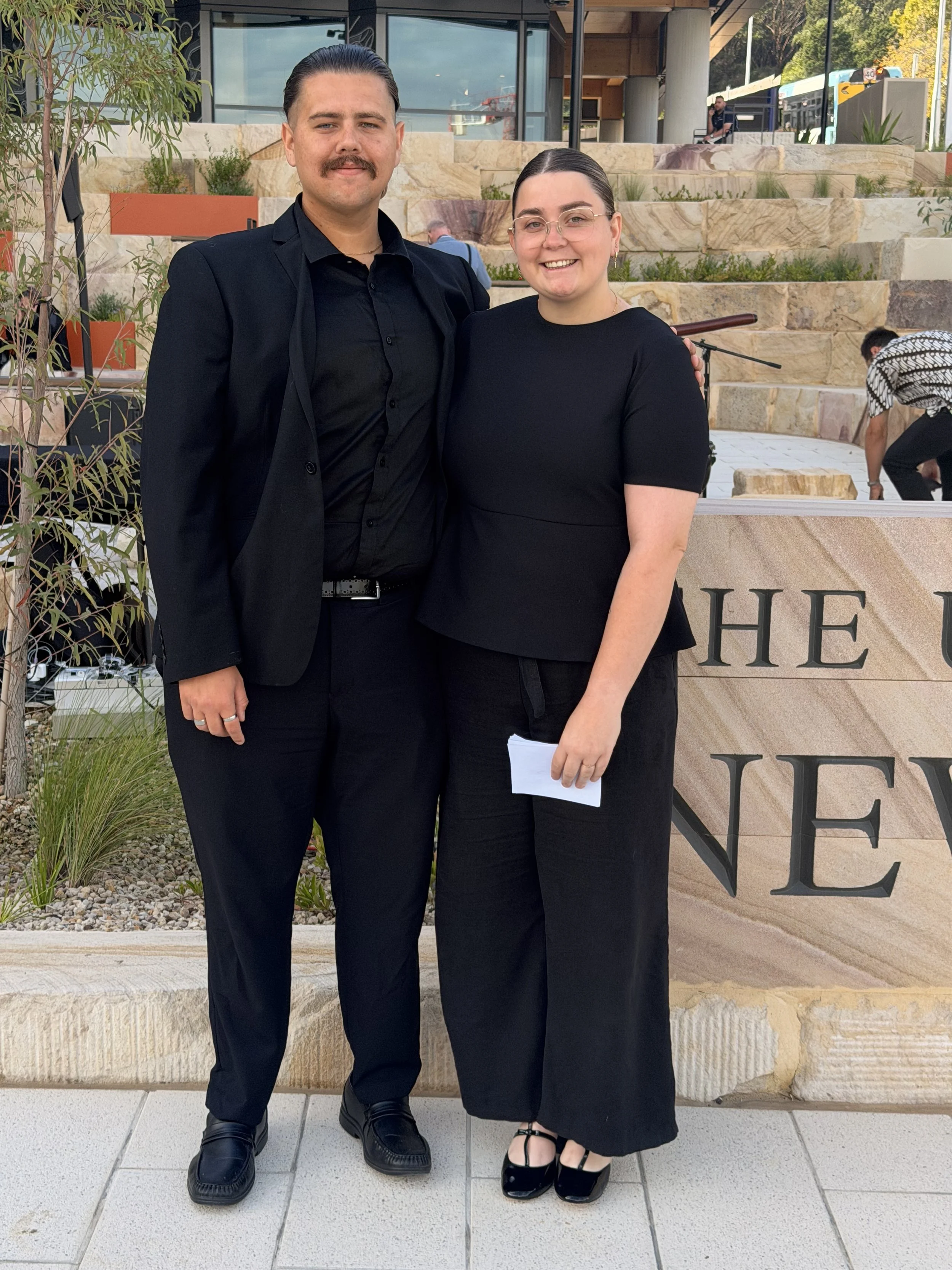 A man and woman dressed in black clothing standing outdoors in front of a stone and glass building, smiling at the camera.