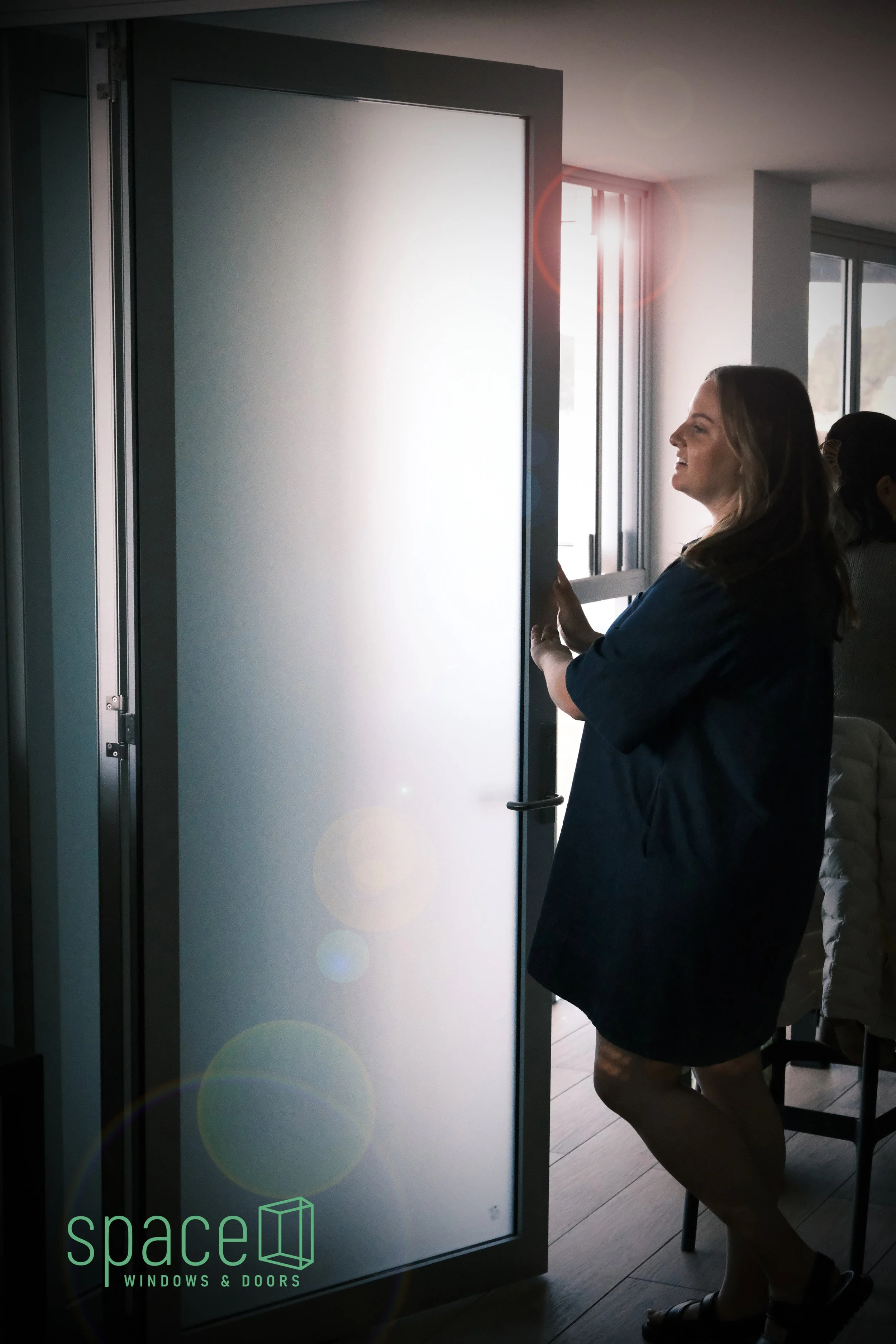 Woman in a blue dress smiles while opening a gray door with a glass panel, in a room with large windows and sunlight streaming in.