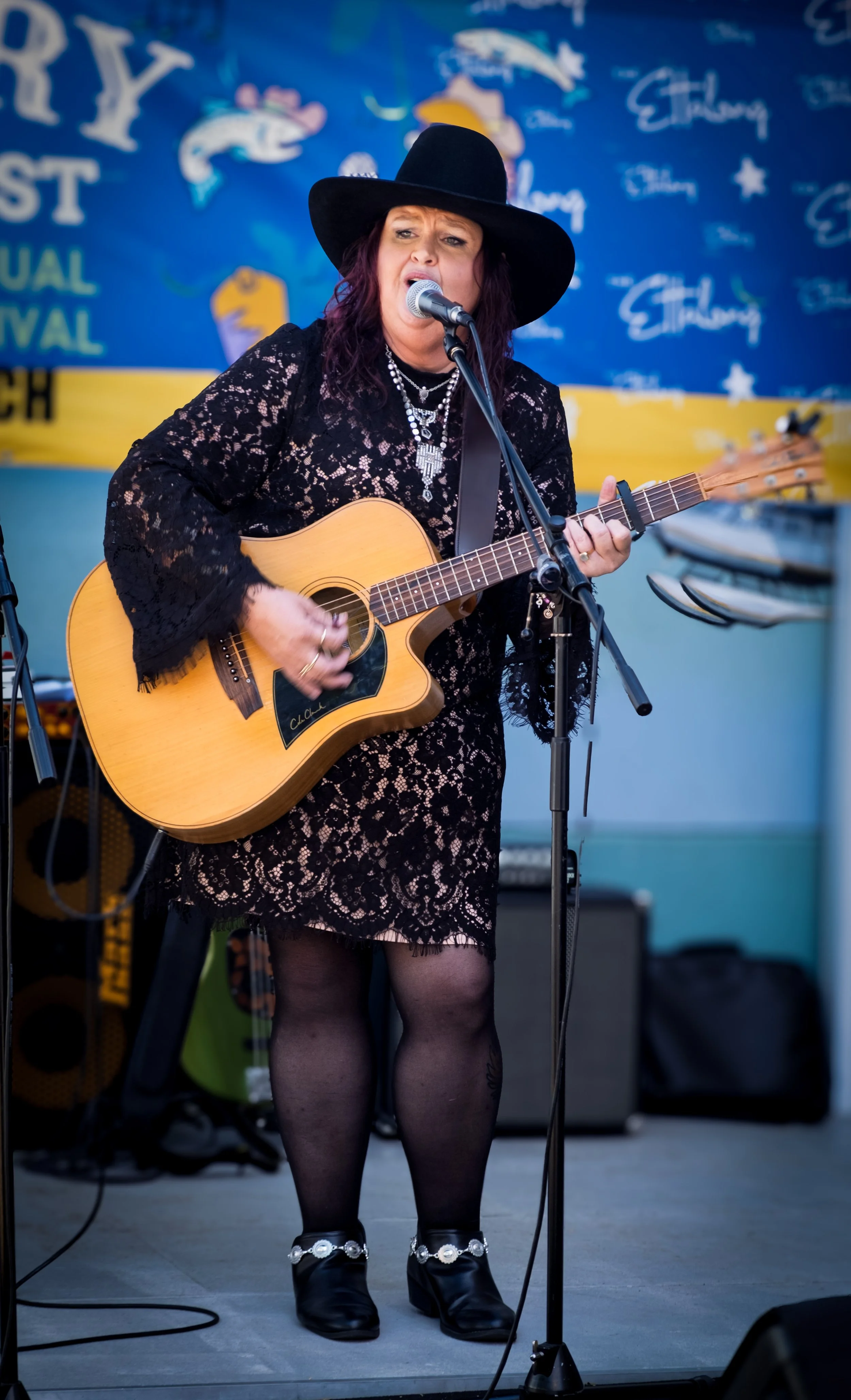 A woman dressed in a black lace dress, black hat, and black boots with decorative silver chains, performing on stage playing an acoustic guitar and singing into a microphone. The background features a colorful festival banner.