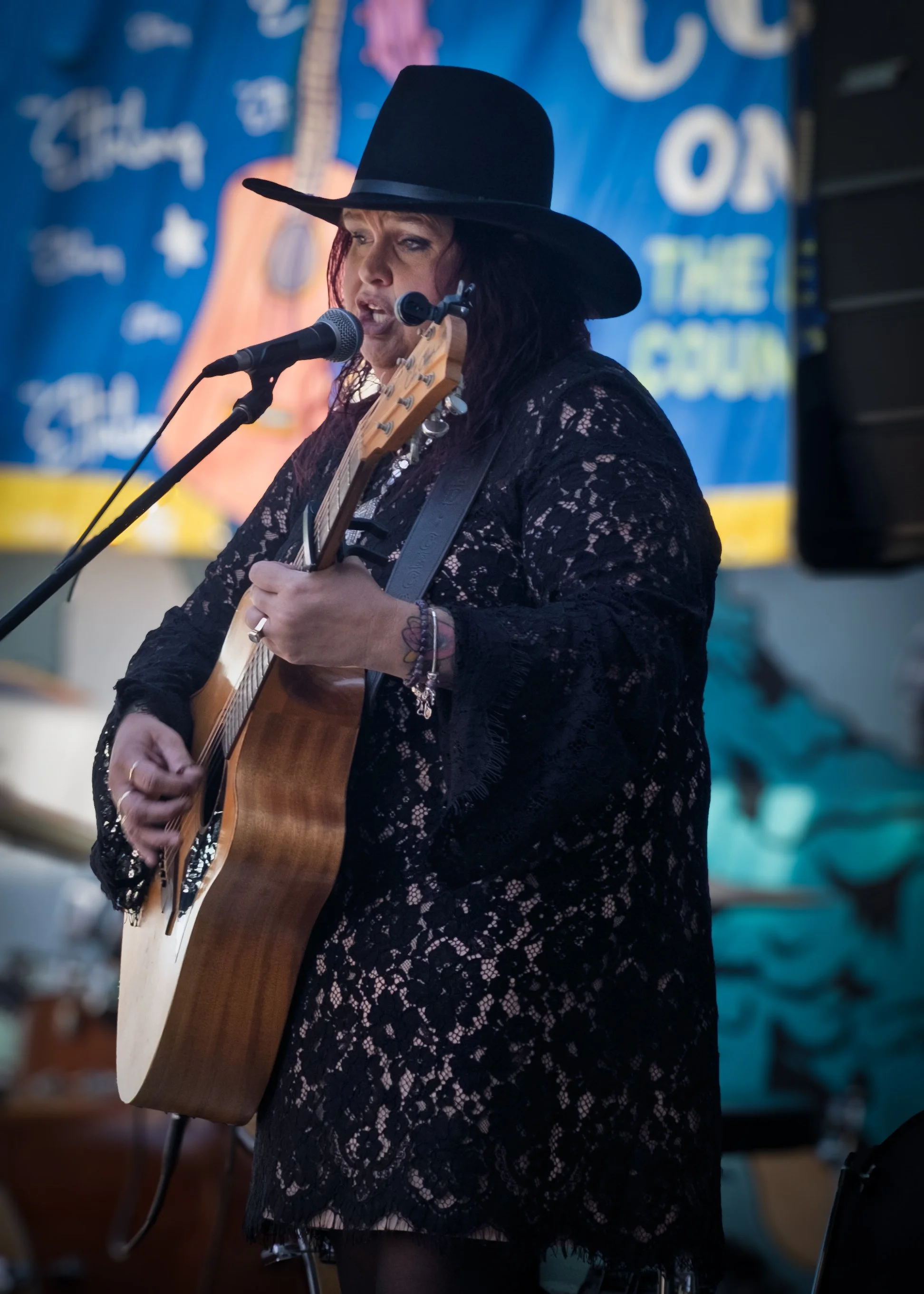 Woman performing guitar on stage, wearing a wide-brimmed black hat and a black lace dress, singing into a microphone.