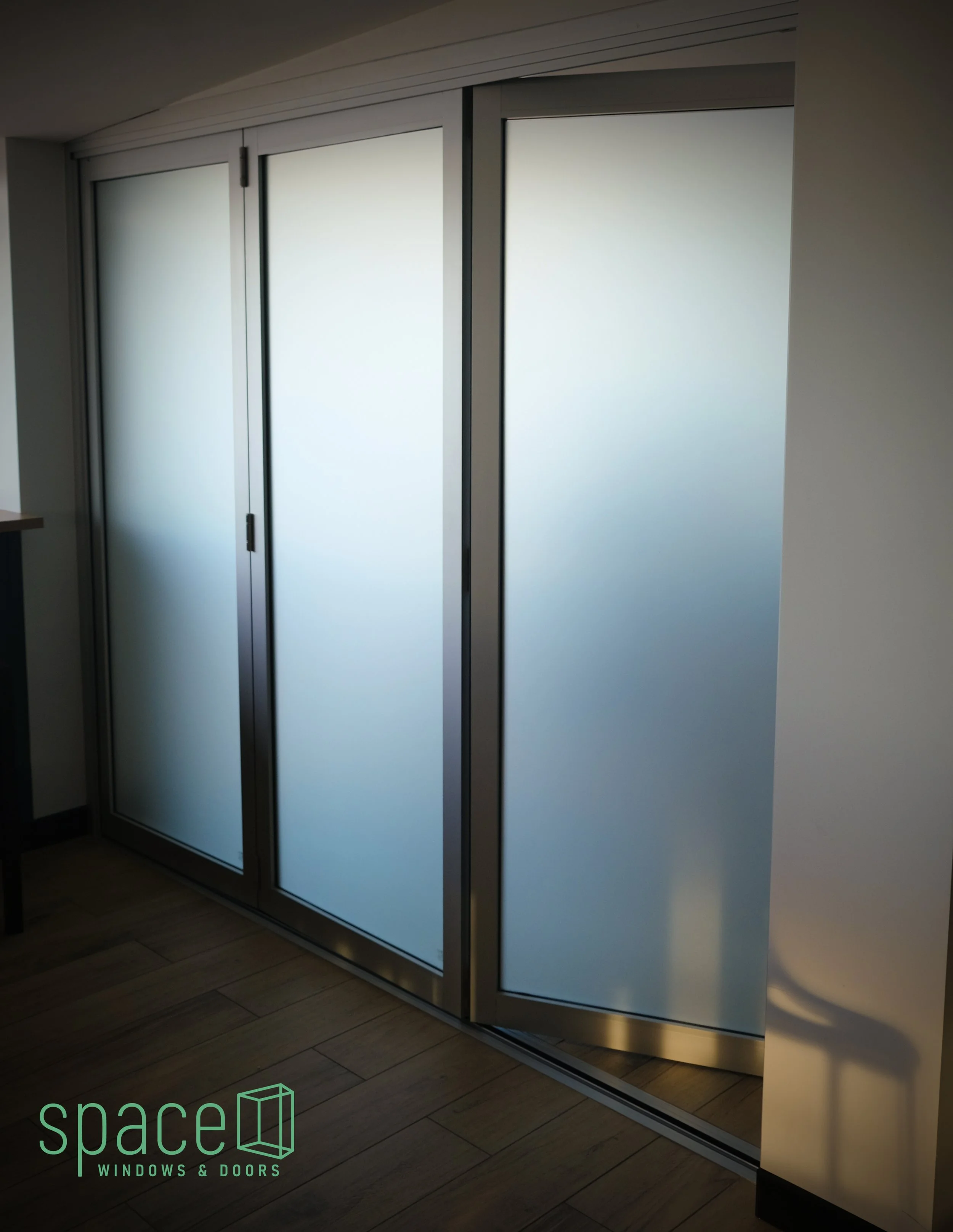 A modern interior with sliding glass doors in silver frames, set in a wall painted in neutral tones. The doors are frosted, allowing light to passage while maintaining privacy. There is a wooden floor and part of a table or counter visible on the left side. The logo of 'space Windows & Doors' is displayed in the bottom left corner.