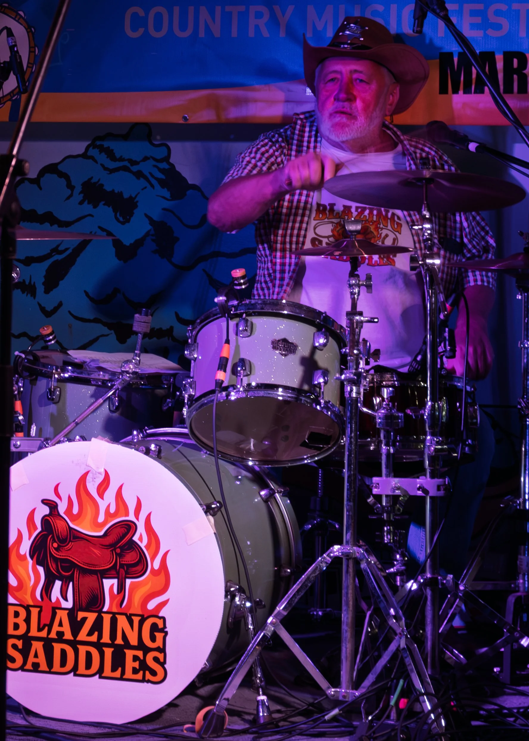 Man playing drums on stage, wearing a cowboy hat, checkered shirt, and a t-shirt with the words 'Blazing Saddles' and a flaming saddle logo, with a blue banner in the background that reads 'Country Music Fest'.