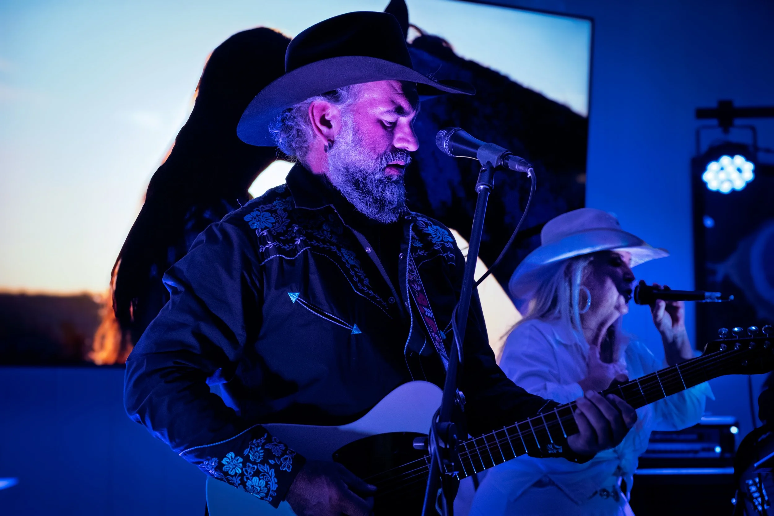 A bearded man wearing a cowboy hat playing an acoustic guitar and singing into a microphone, with a woman also singing and playing guitar beside him, during a live performance at sunset or in low lighting.