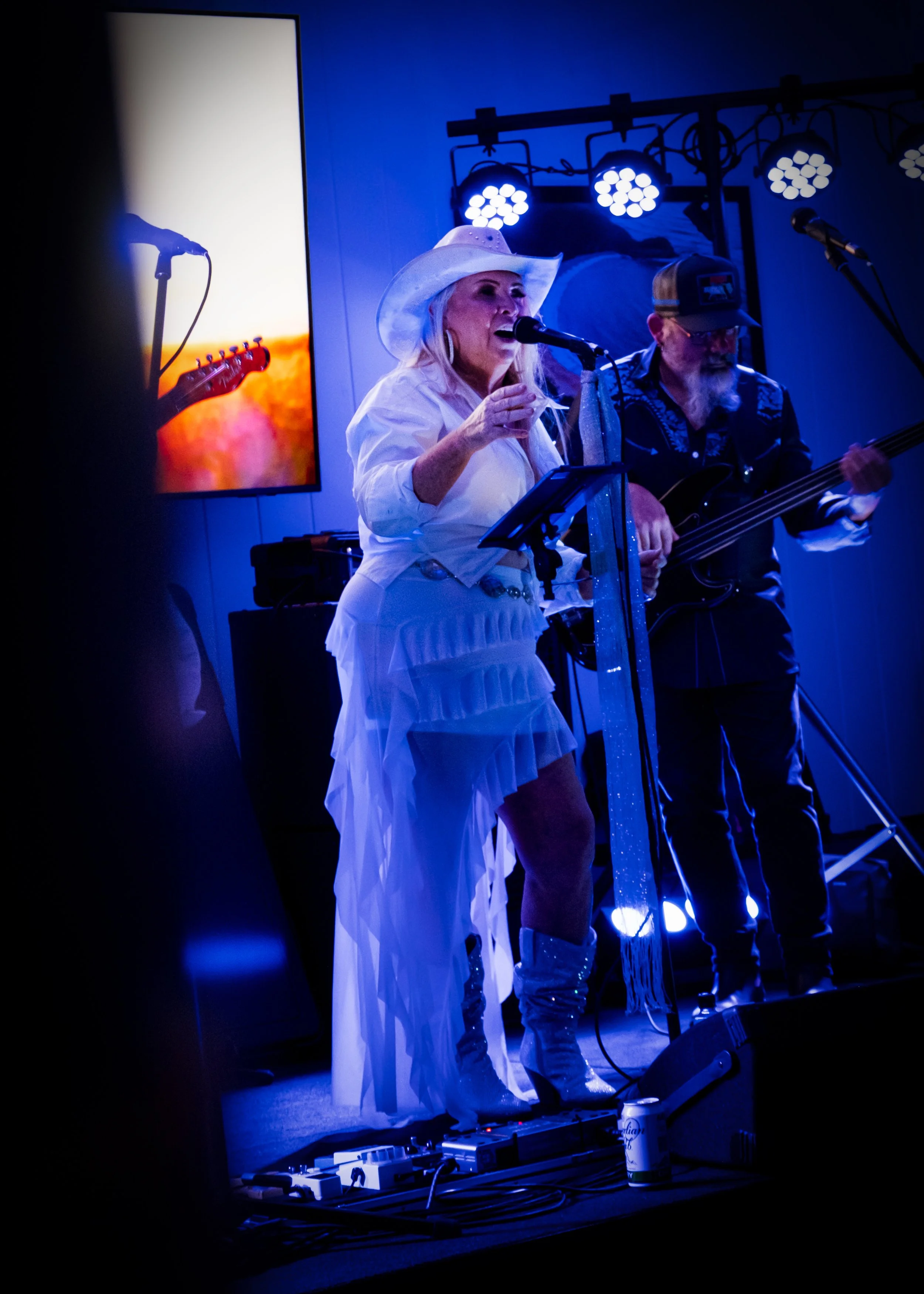 An elderly woman singing into a microphone on stage with a man playing guitar next to her, illuminated by stage lights; a large screen displays a sunset background with a silhouette of a microphone.
