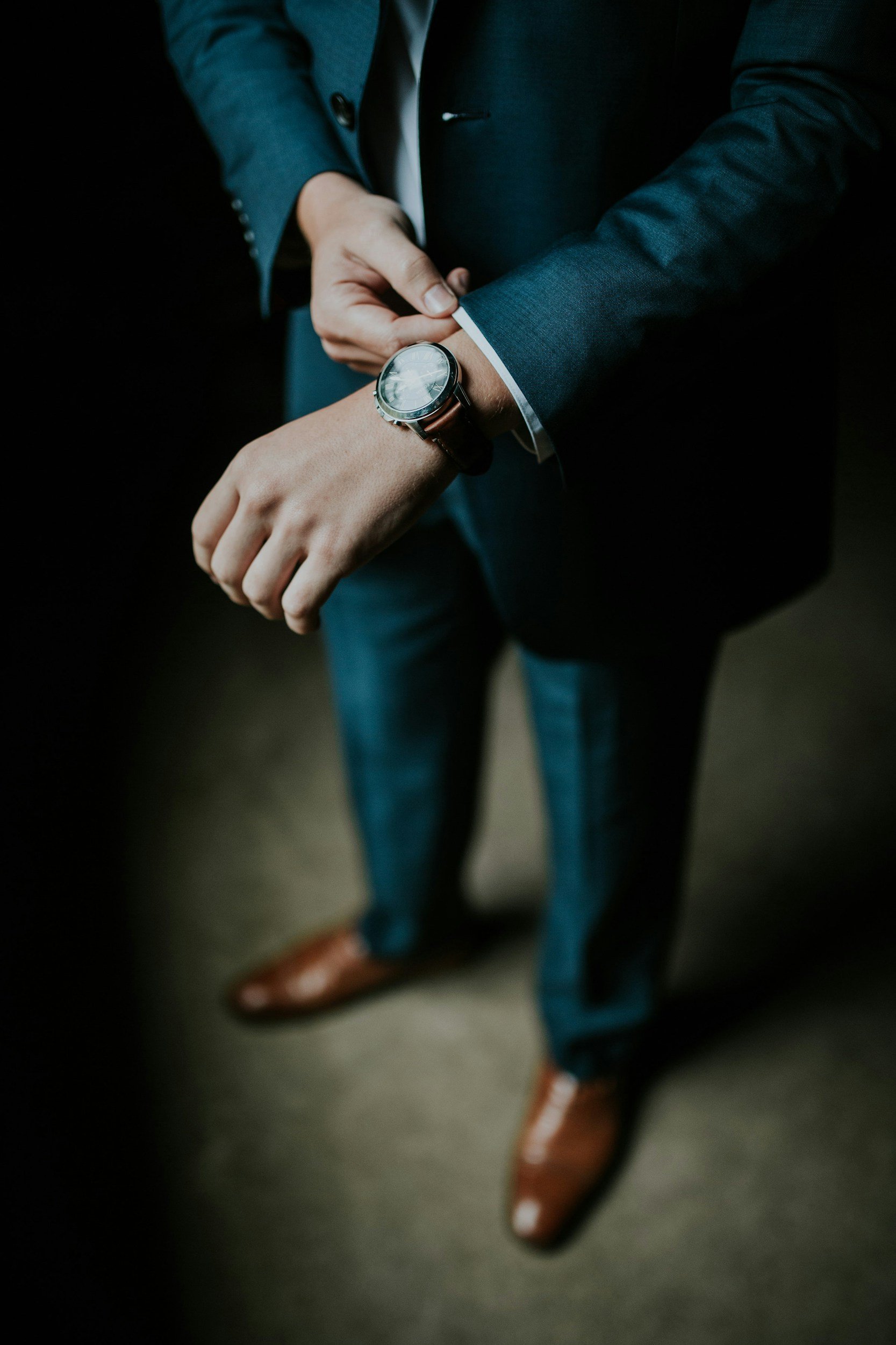 Person in a dark suit adjusting their wristwatch, wearing brown leather shoes and blue trousers.