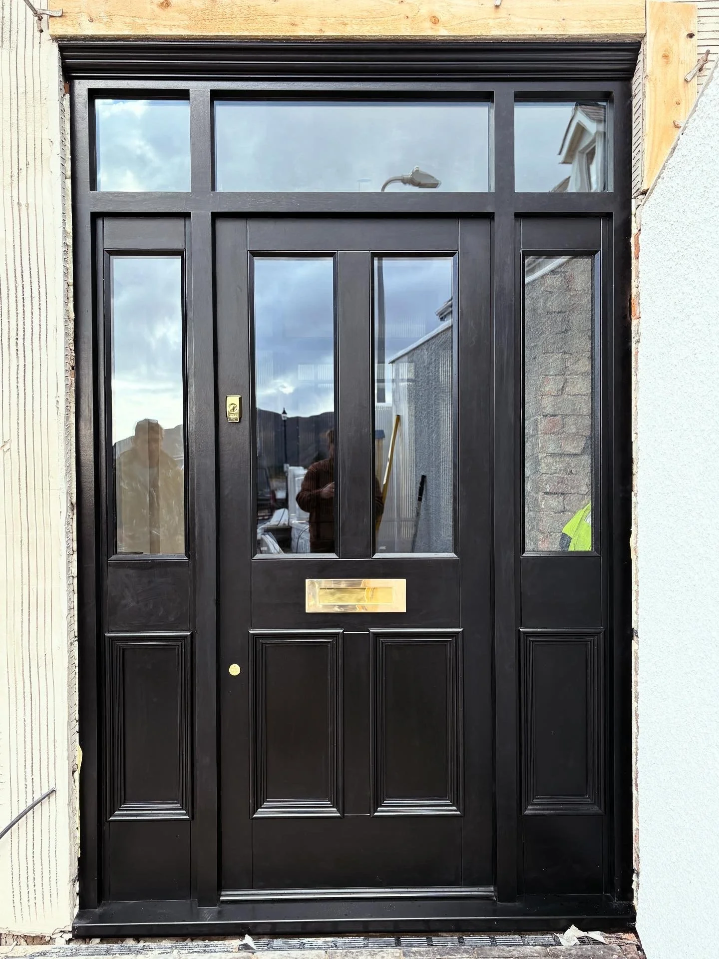 Front door with black wooden panels, glass window panes at the top, brass mail slot, and a brass doorknob, set in a partially constructed brick and wood frame. in Colwyn heights 