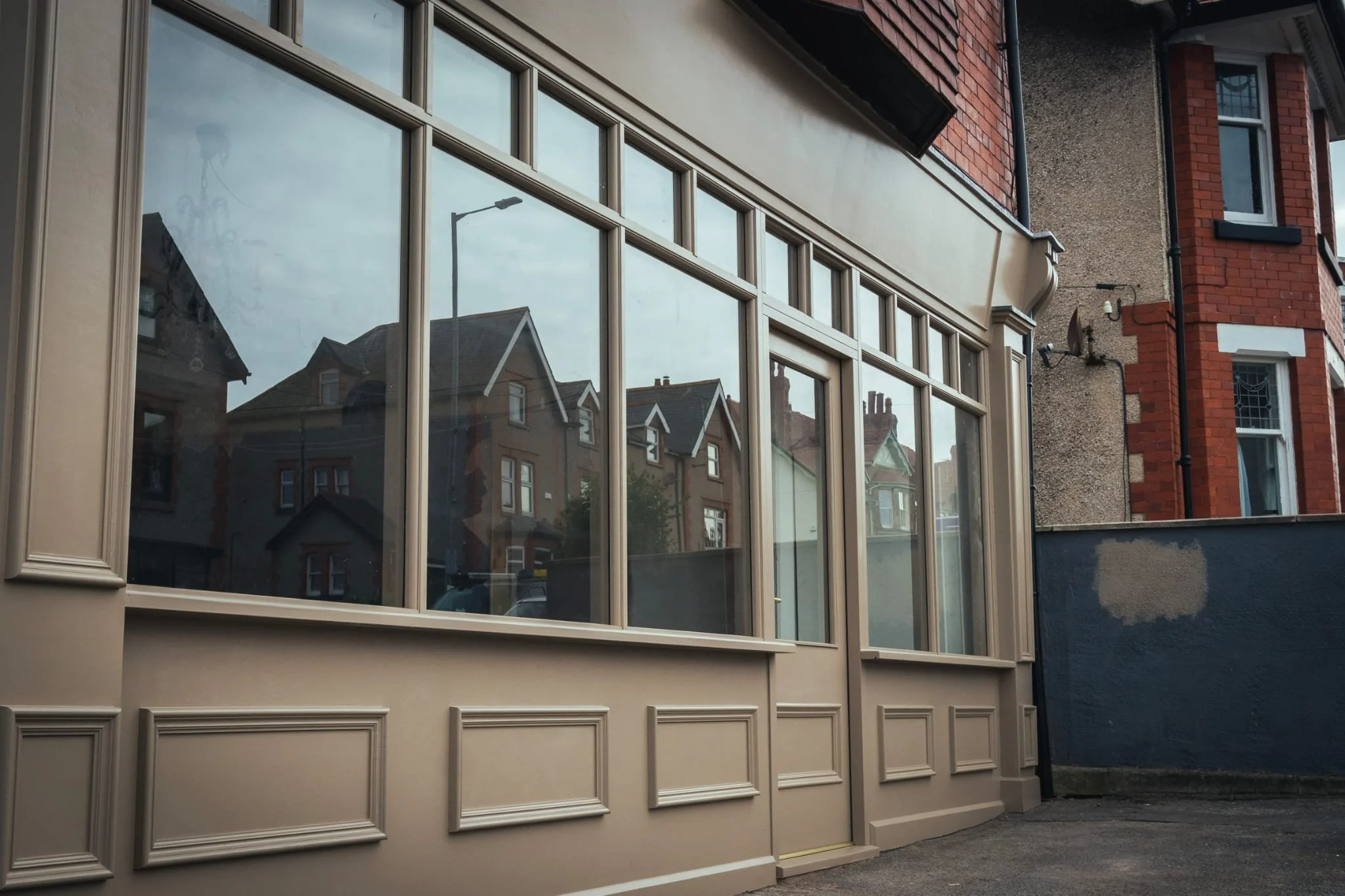 Side view of a building with large windows and decorative panels below, reflected houses and a streetlamp in the glass, with a brick and concrete building nearby.