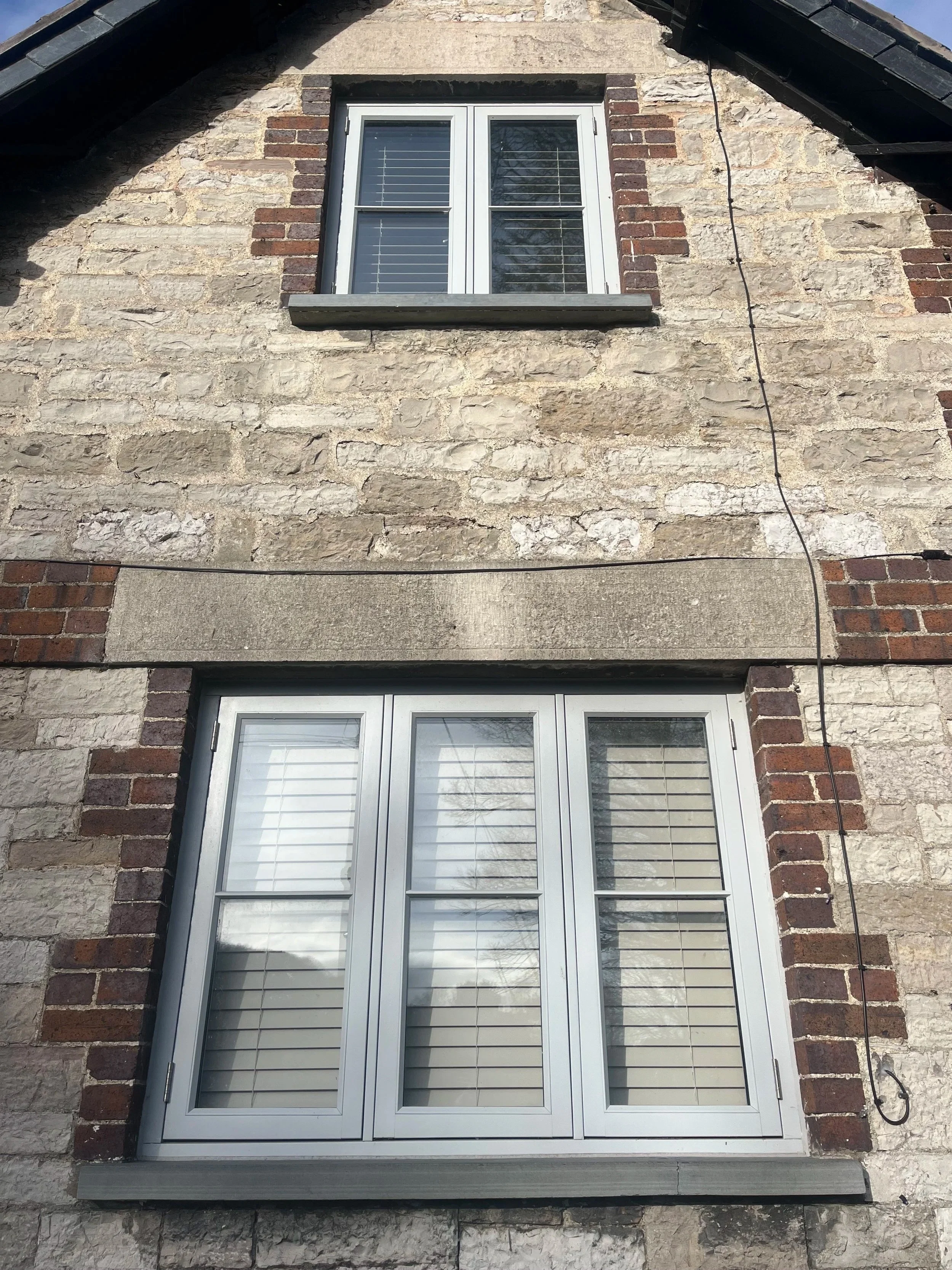 A close-up of a stone house wall with a white-framed window and white shutters, with brick accents around the window and a black cable running along the right side.