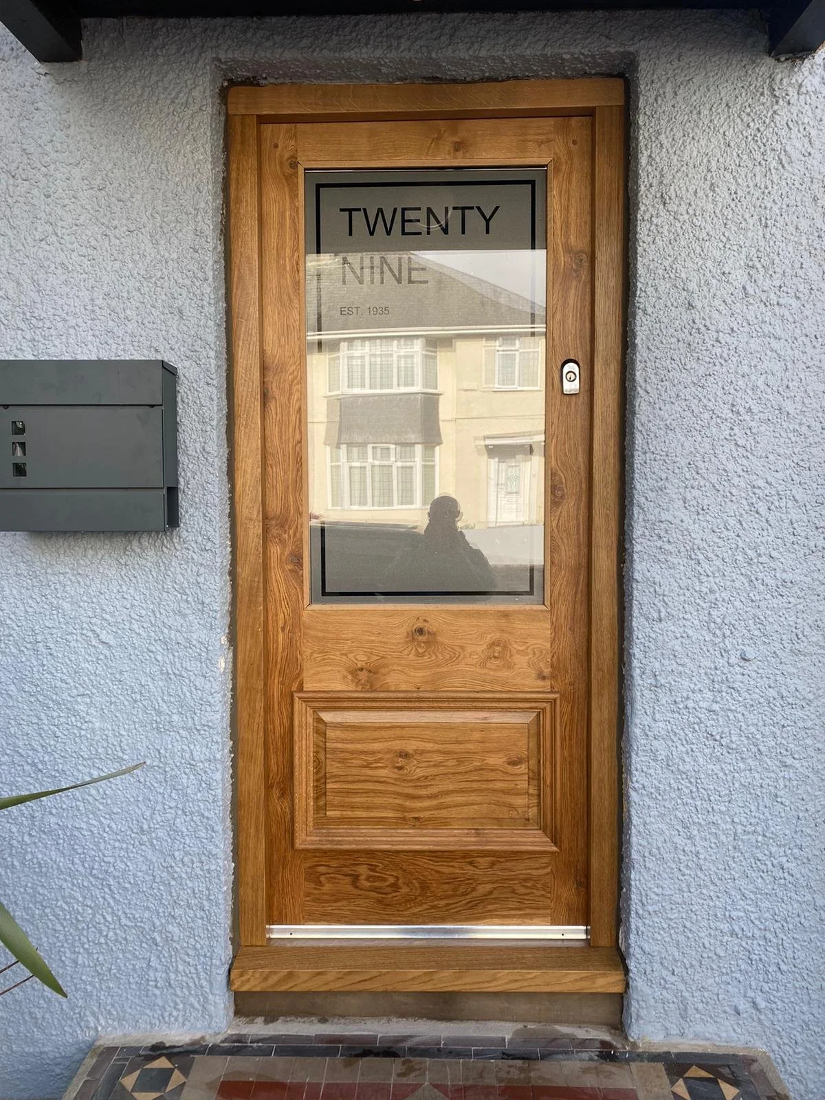 Front door with a glass window displaying the words "TWENTY NINE EST. 1935" in black lettering, surrounded by a wooden frame, with a gray textured wall and a mailbox to the left. made for a customer in London 
